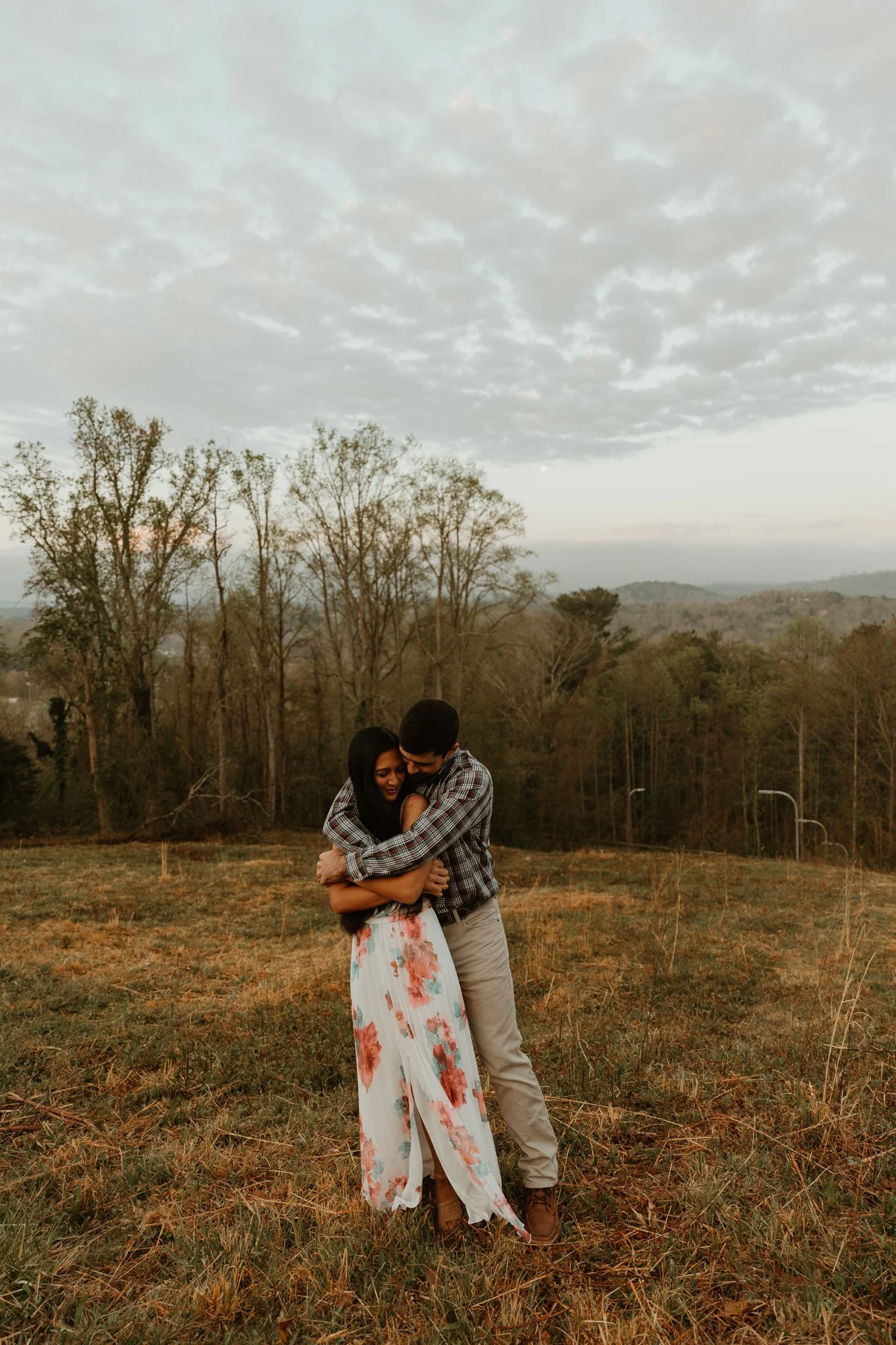 A couple hugging in a field with trees and hills in the background during dusk, cloudy sky overhead.