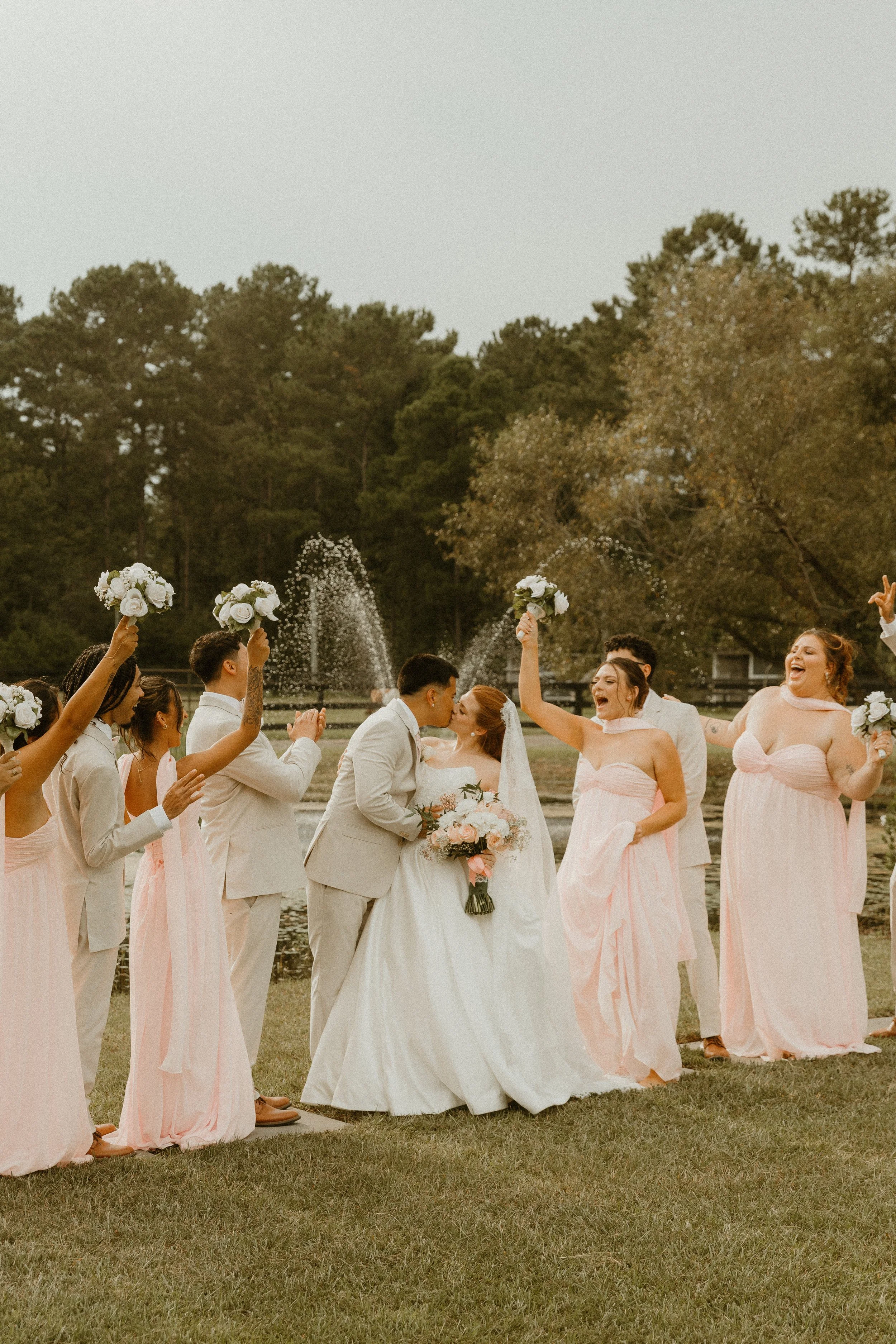 A wedding celebration outdoors with a bride and groom kissing surrounded by bridesmaids and groomsmen, people holding bouquets and throwing petals or confetti, and a fountain in the background.