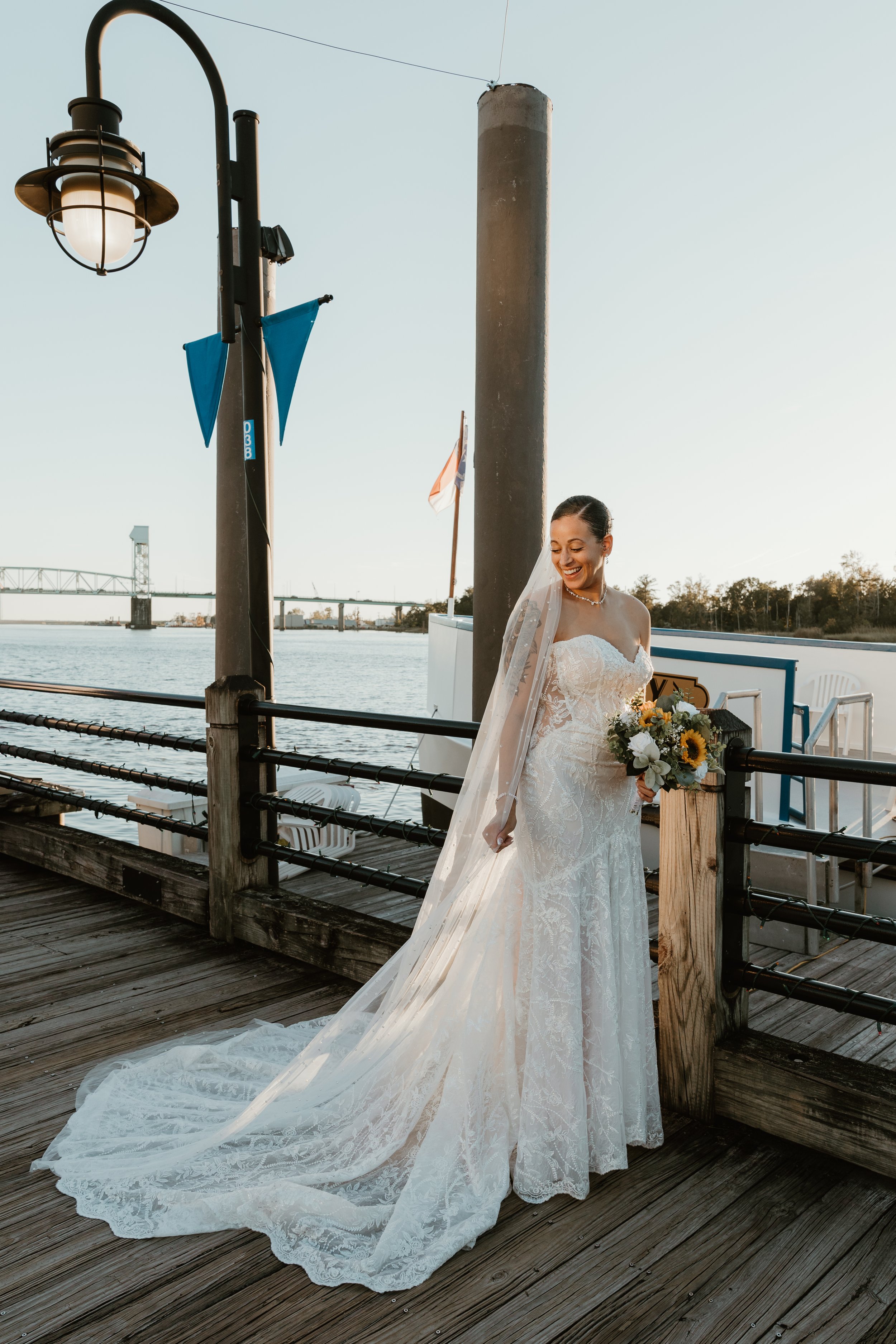 A bride in a white lace wedding gown with a long train standing on a wooden dock near the water, holding a bouquet of sunflowers and white flowers, smiling with her eyes closed, during sunset.