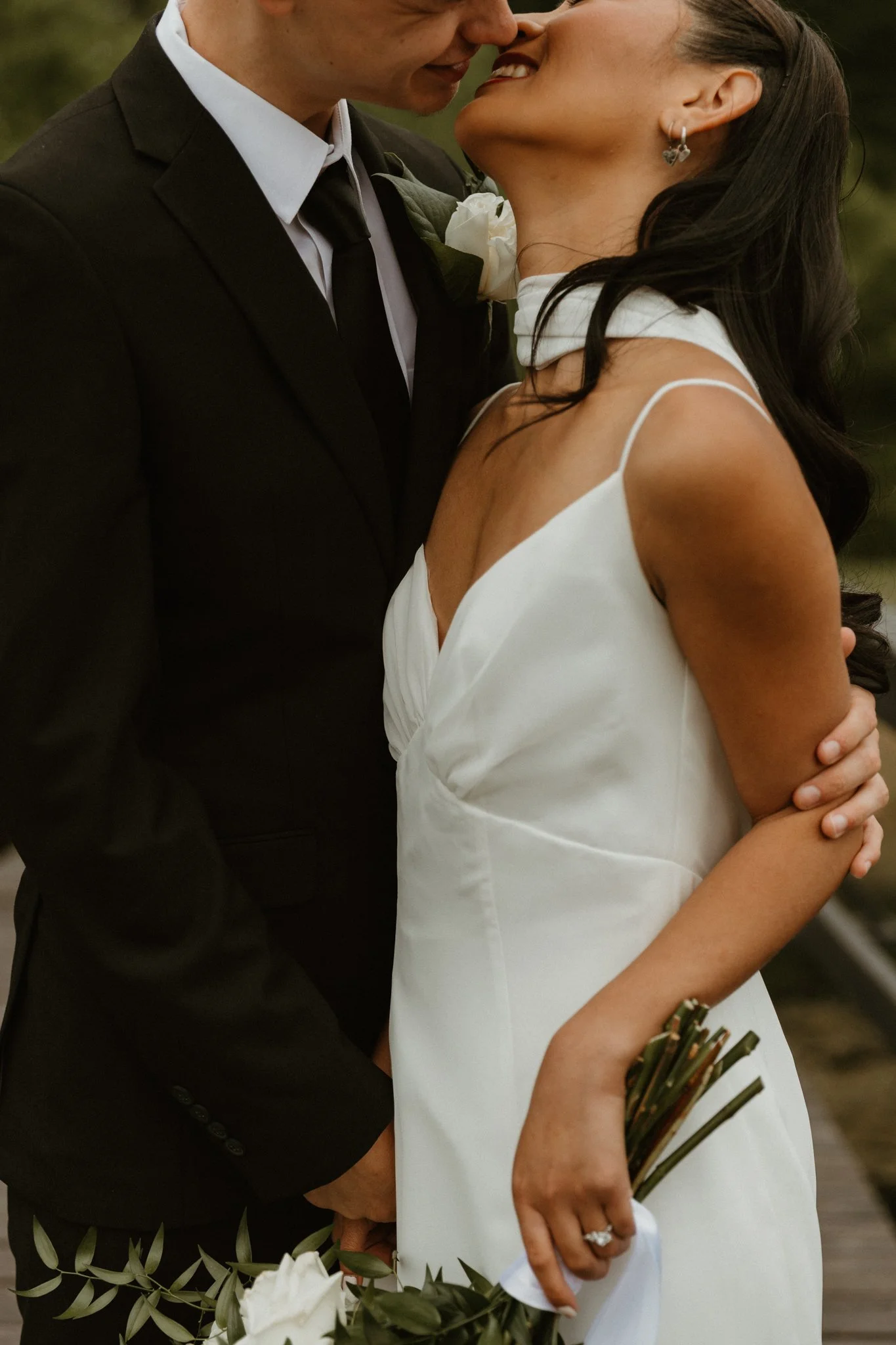 A couple dressed in wedding attire sharing a close, intimate moment, with the man in a black suit and the woman in a white dress, holding a bouquet of flowers.