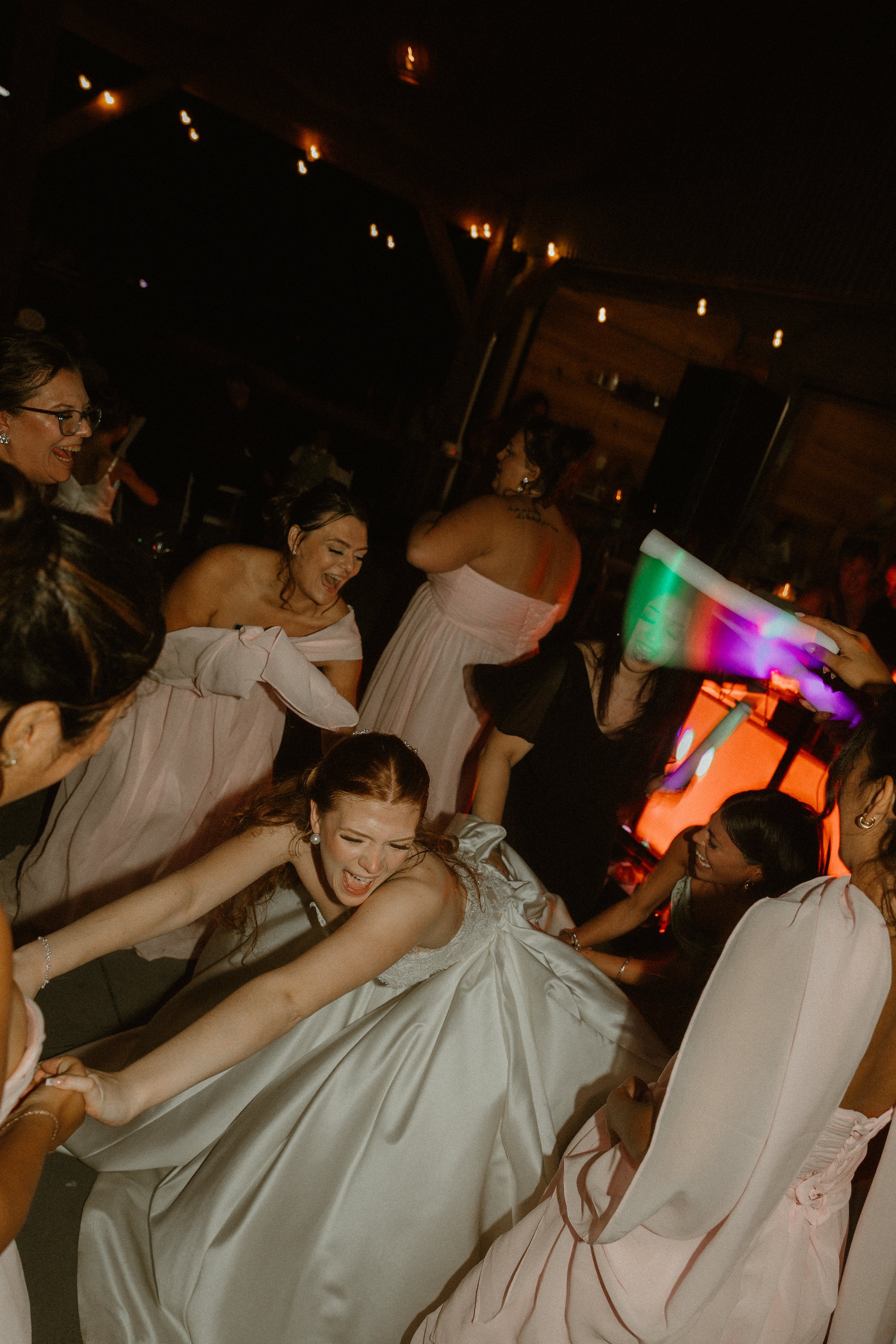 A group of women in a celebration, with some wearing fancy dresses, dancing and having fun indoors at night, with dim lighting and a wooden interior.