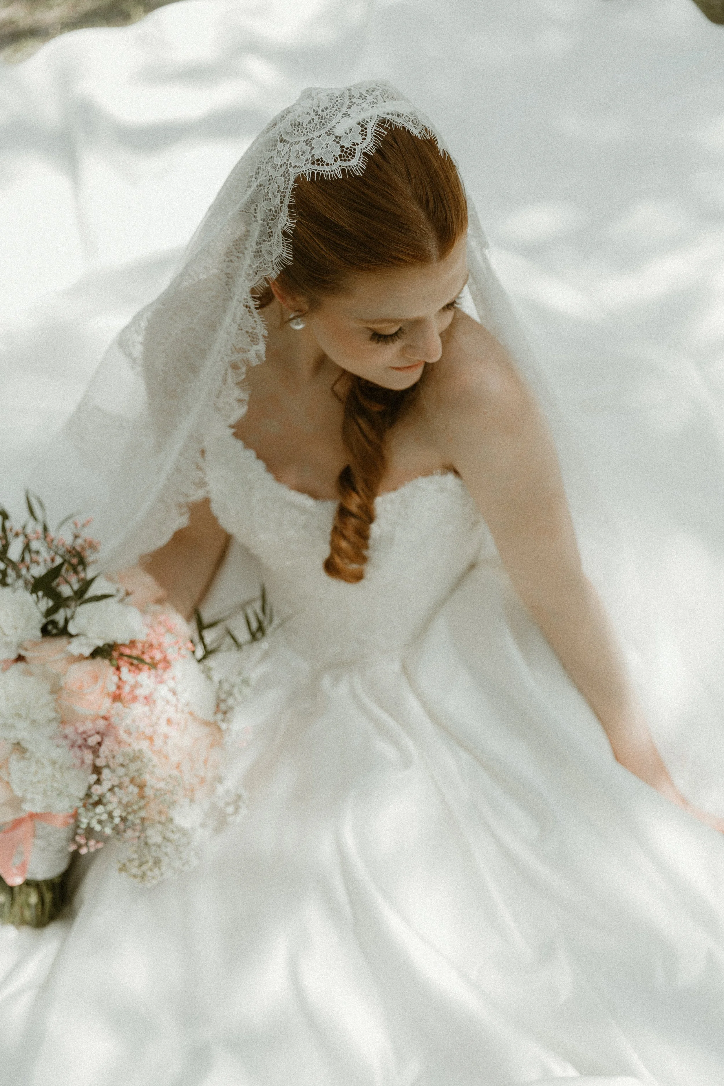 A bride with red hair and a lace veil sitting on a white satin wedding dress, holding a bouquet of pink and white flowers.
