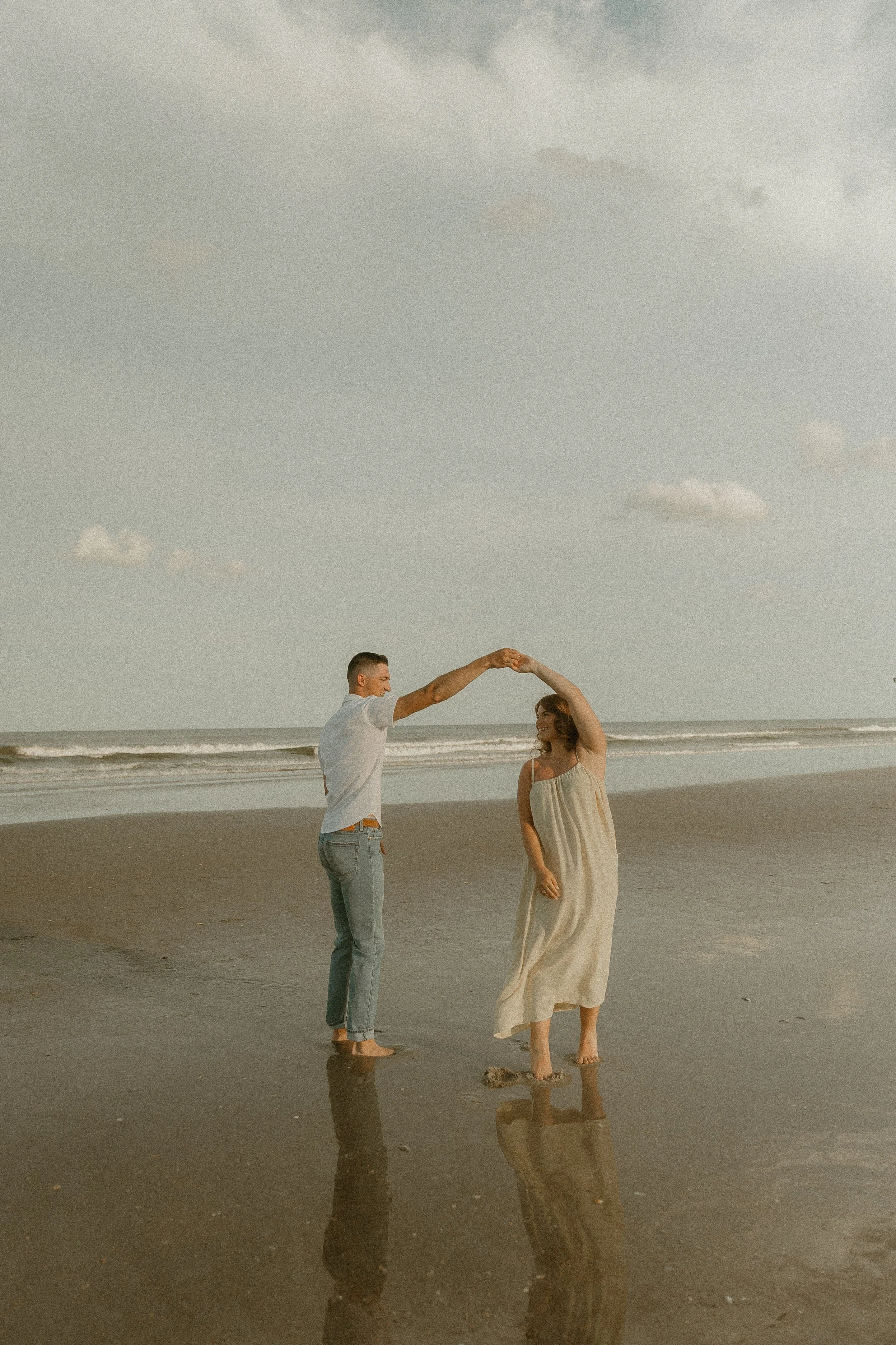 A couple dancing on the beach at sunset, with the man in a white shirt and jeans, and the woman in a light dress, holding hands and looking at each other, their reflection visible on the wet sand.