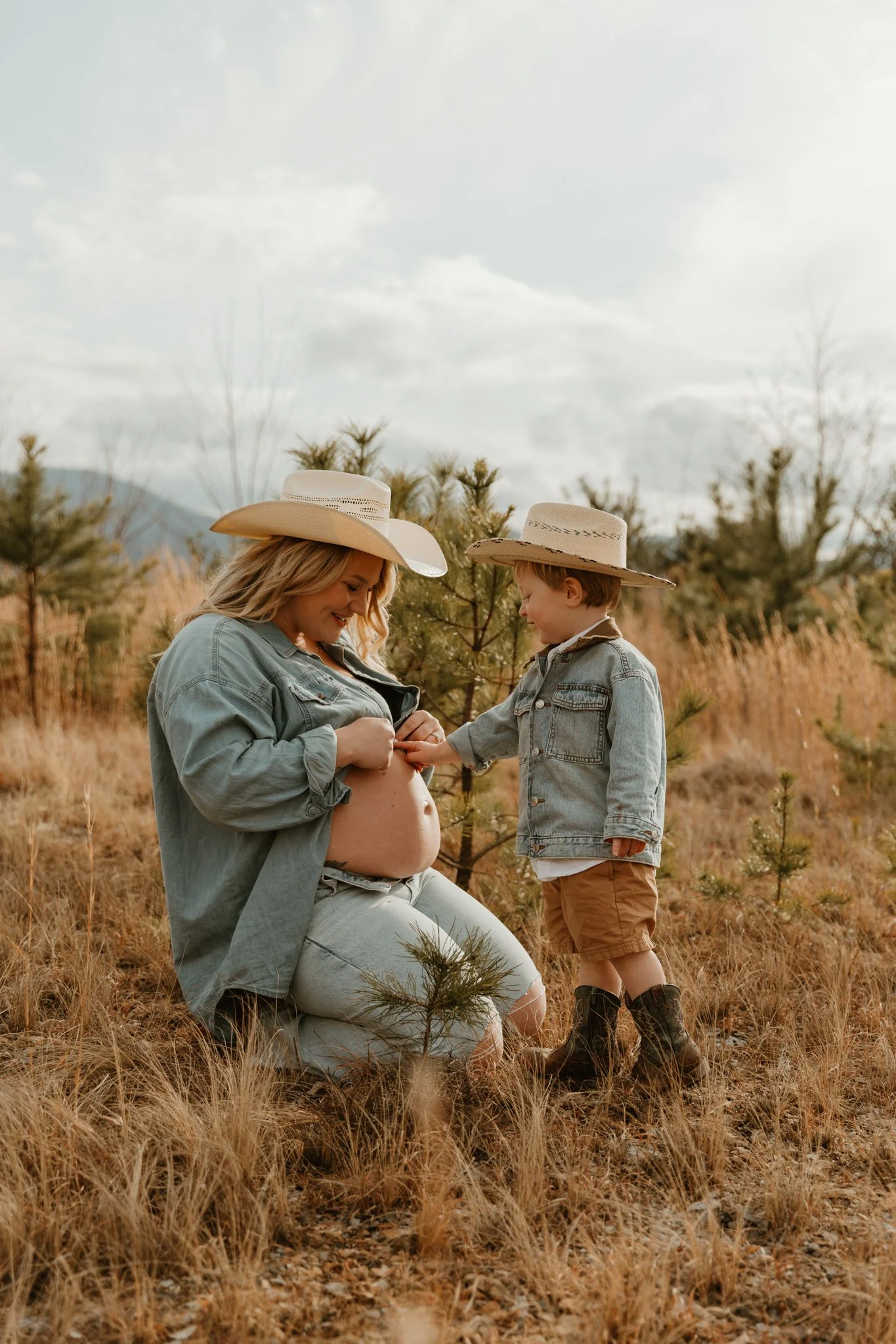 A pregnant woman and her young son outdoors on a grassy field, both wearing cowboy hats and denim jackets. The boy is touching the woman's belly.