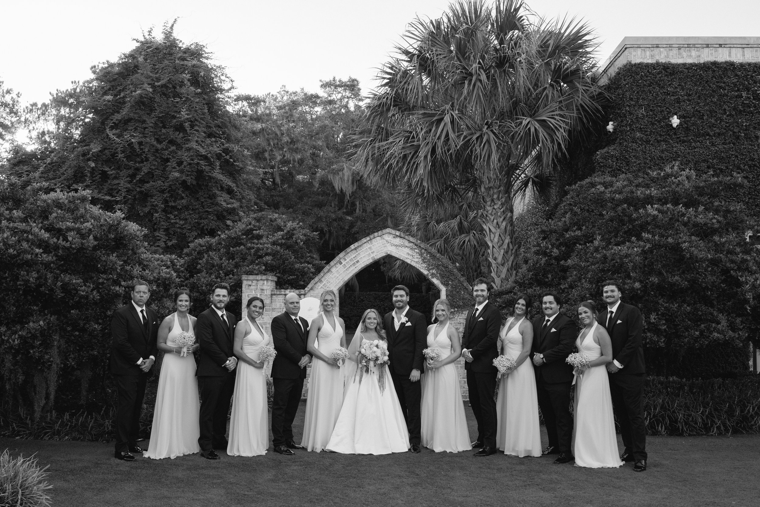 Black and white photograph of a wedding party with bridesmaids in long dresses and groomsmen in suits, standing outdoors under a large palm tree and other trees, with an archway and building in the background.