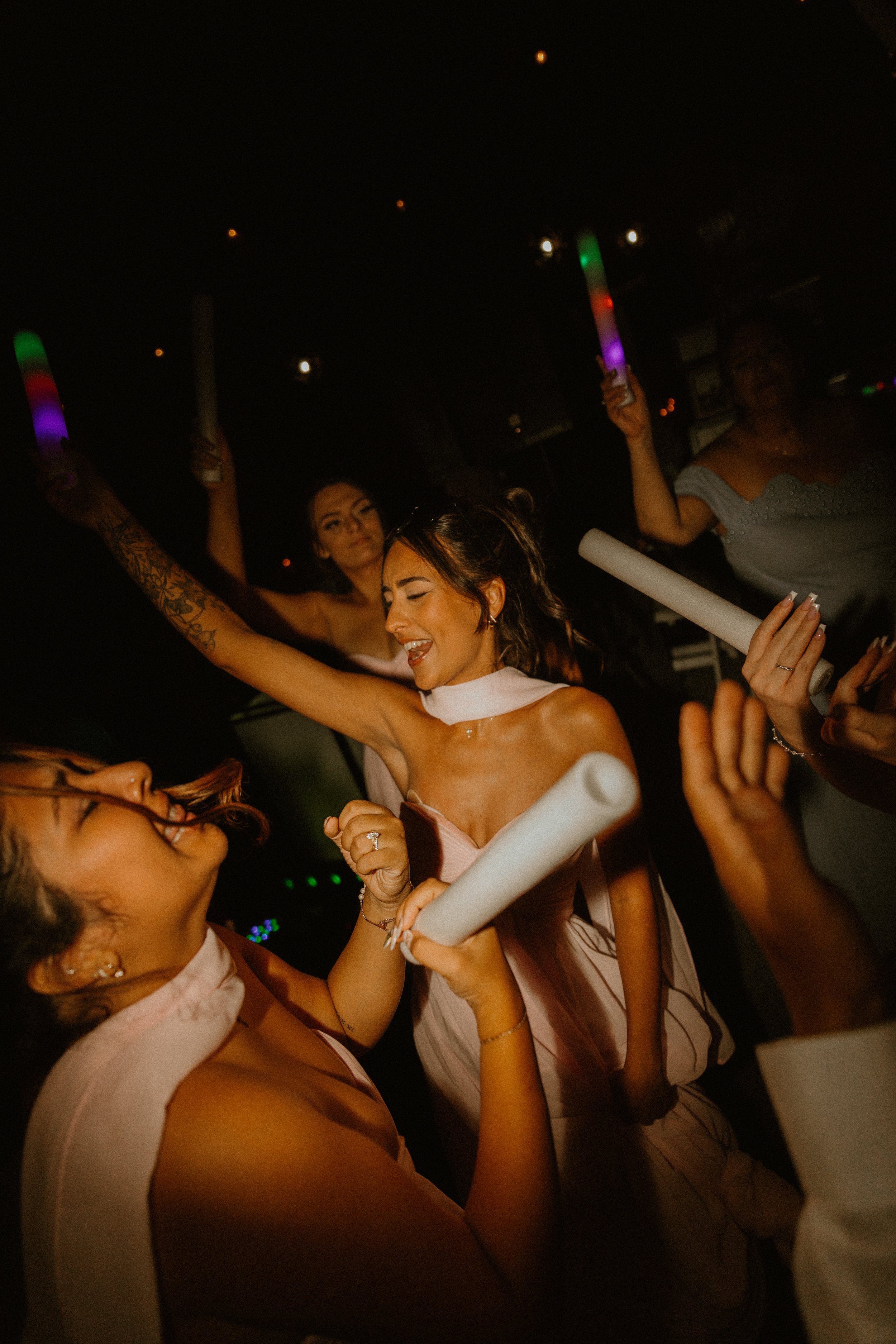 Group of women celebrating at a party, holding glow sticks and wearing elegant attire, with one woman dancing and smiling.