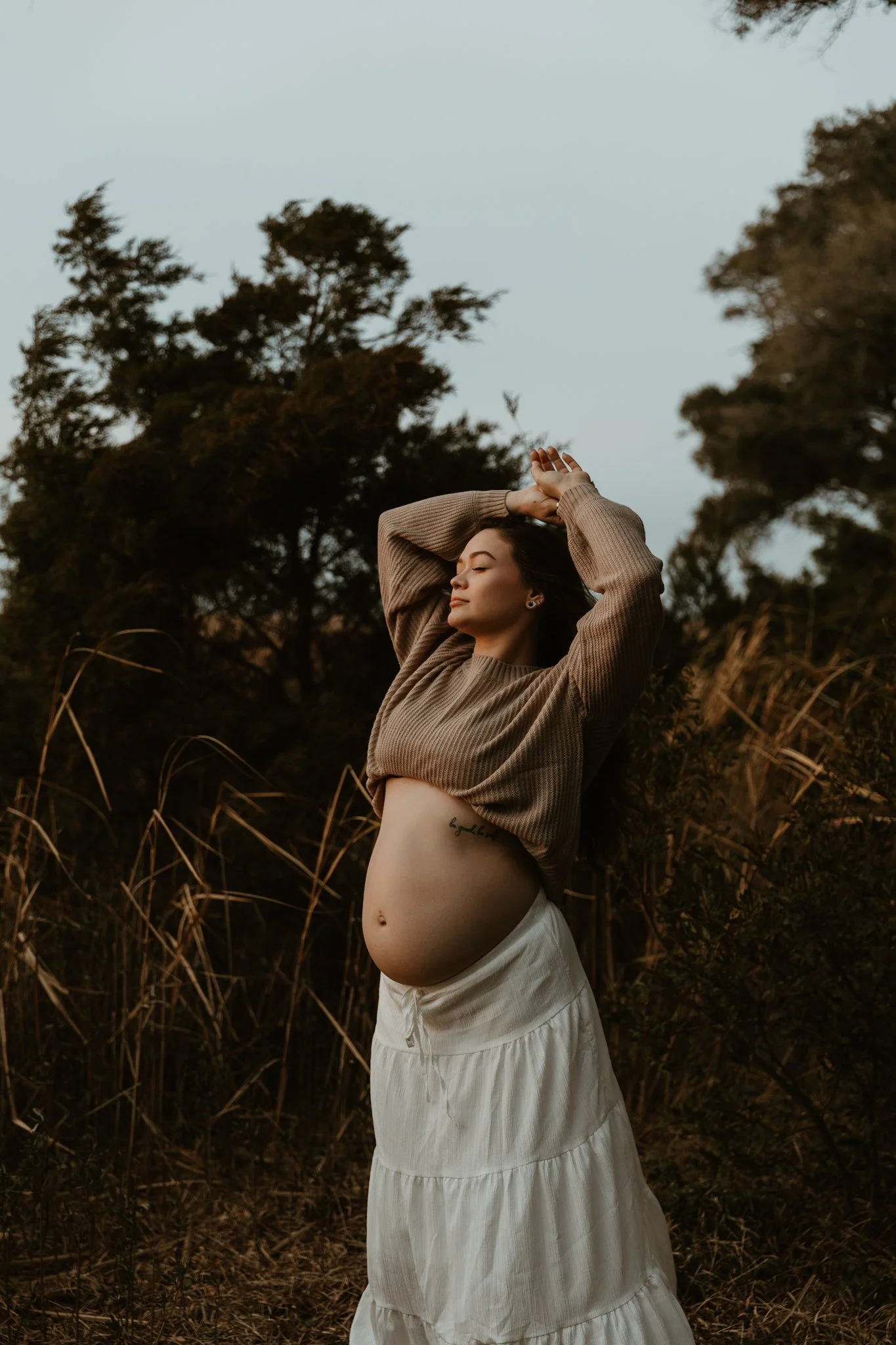 A pregnant woman standing outdoors with her arms raised above her head, eyes closed, and a peaceful expression on her face.