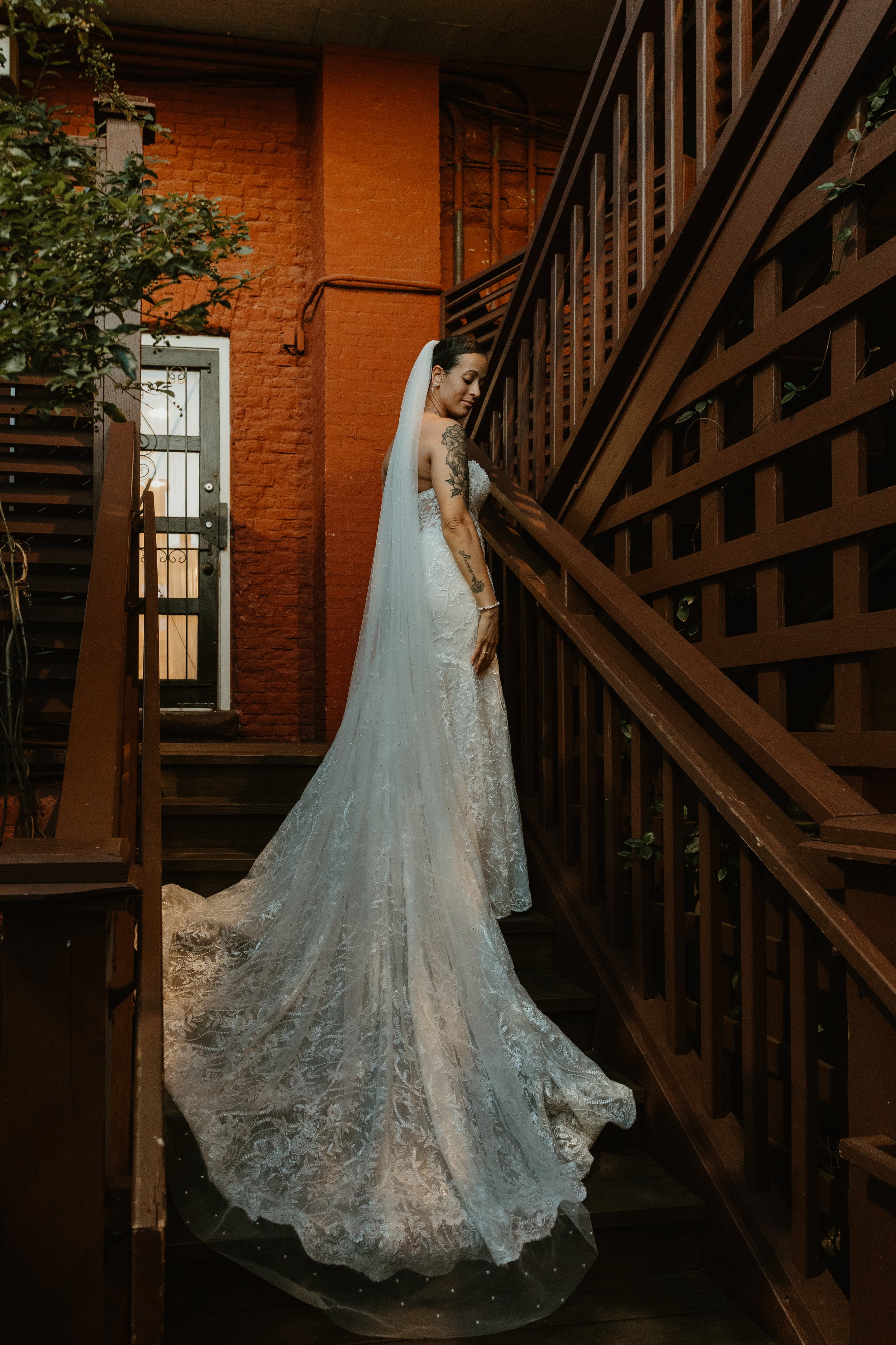 Bride in a white lace wedding gown and veil standing on stairs, indoor setting with brick wall and wooden handrails.