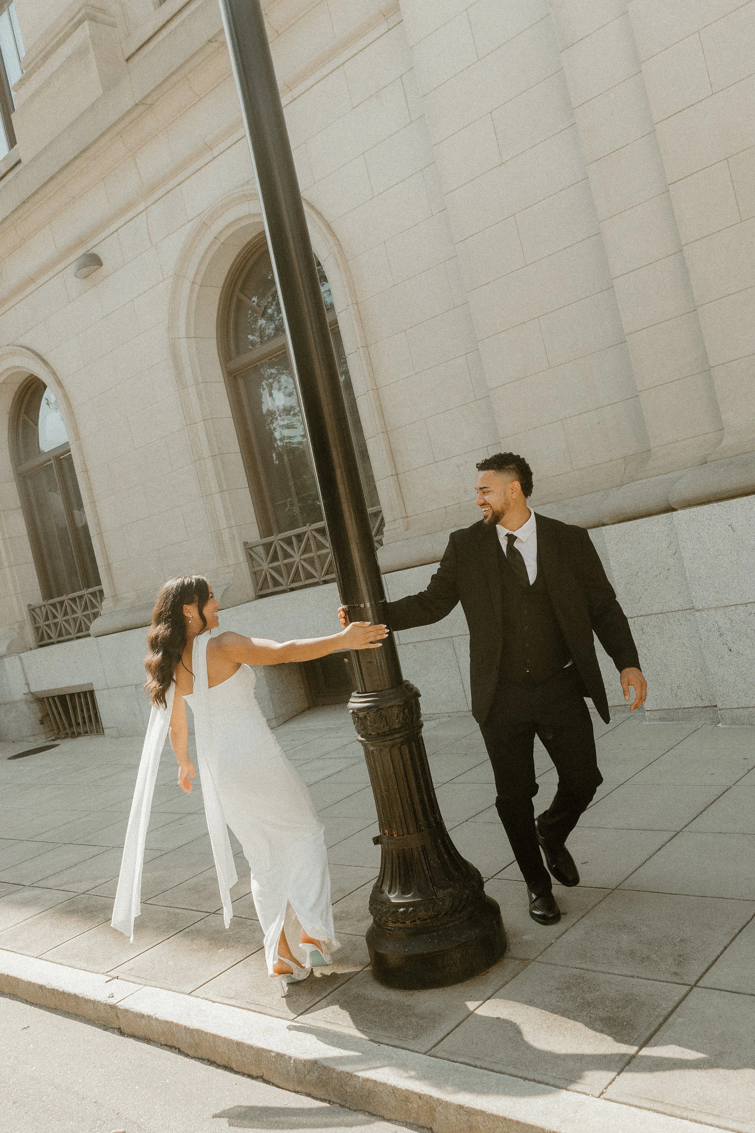 A bride in a white wedding dress reaching out her hand for a handshake with a groom in a black suit and tie, under a streetlamp on a city sidewalk.