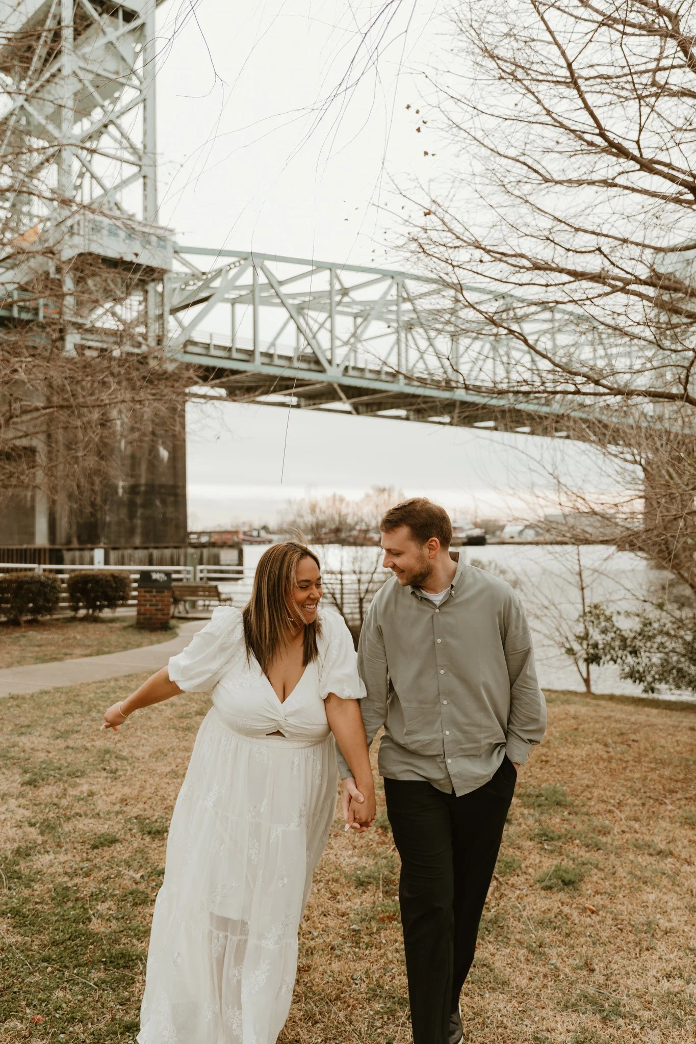 A smiling couple holding hands and walking outdoors near a river, with leafless trees and a bridge in the background.