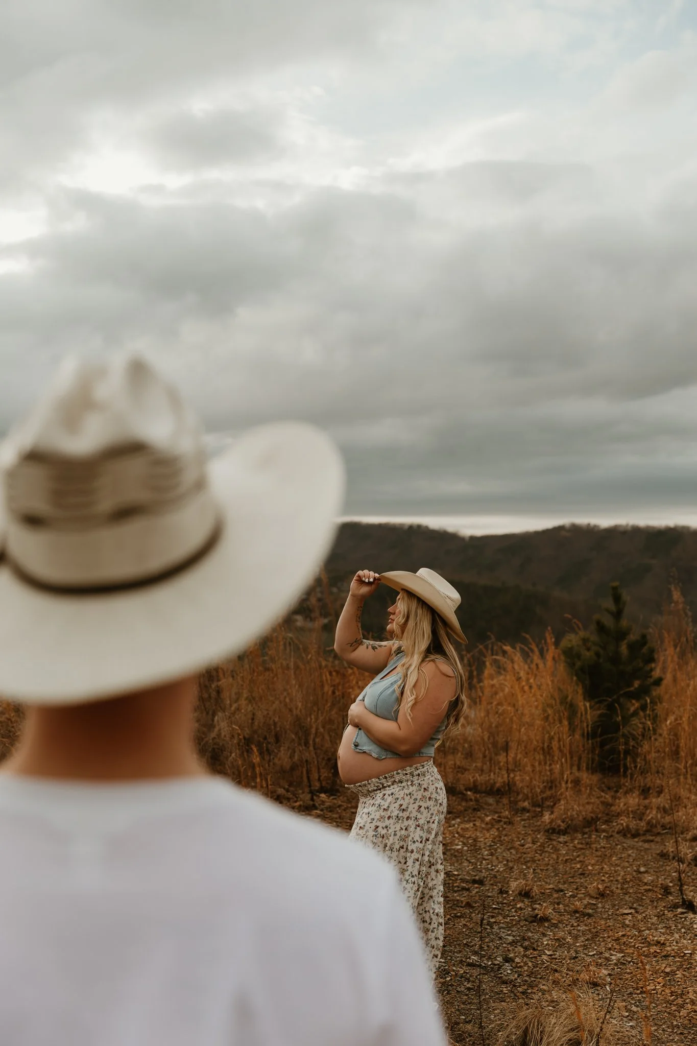 A pregnant woman with long blonde hair, wearing a light blue top, floral skirt, and cowboy hat, standing outdoors on a dirt path with dry grass and trees, holding her belly with one hand and touching her hat with the other, as a person in the foregro