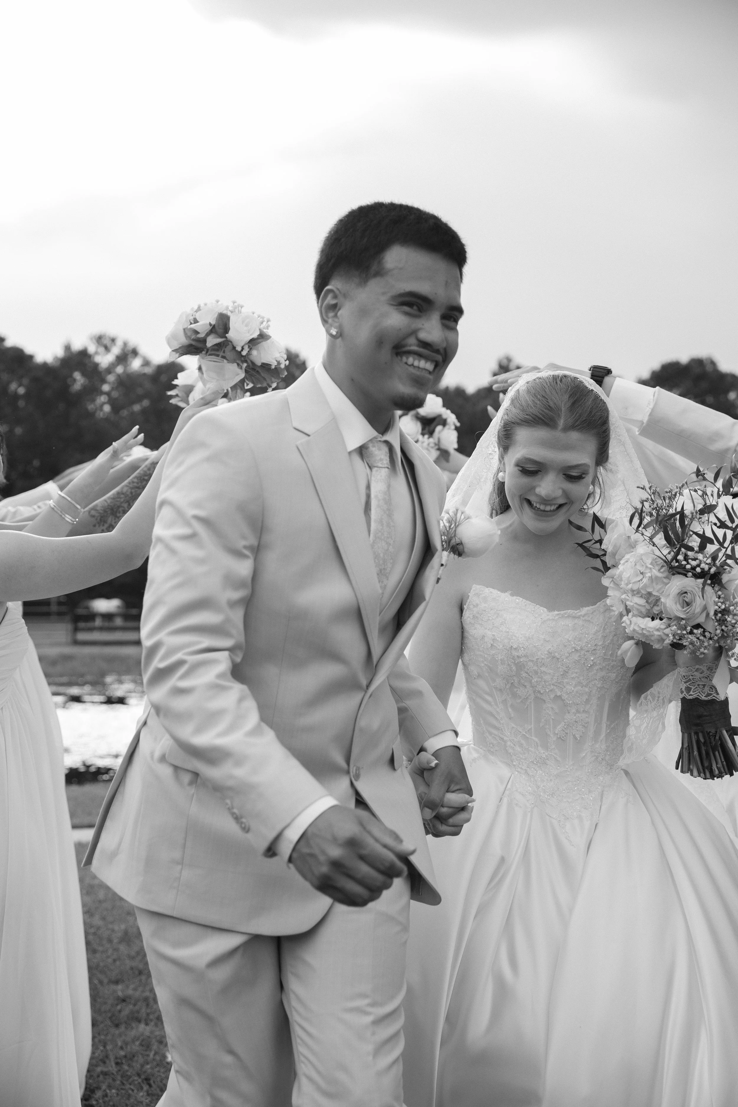 A black-and-white photo of a bride and groom holding hands, smiling, surrounded by bridesmaids holding bouquets, outdoors with trees in the background.