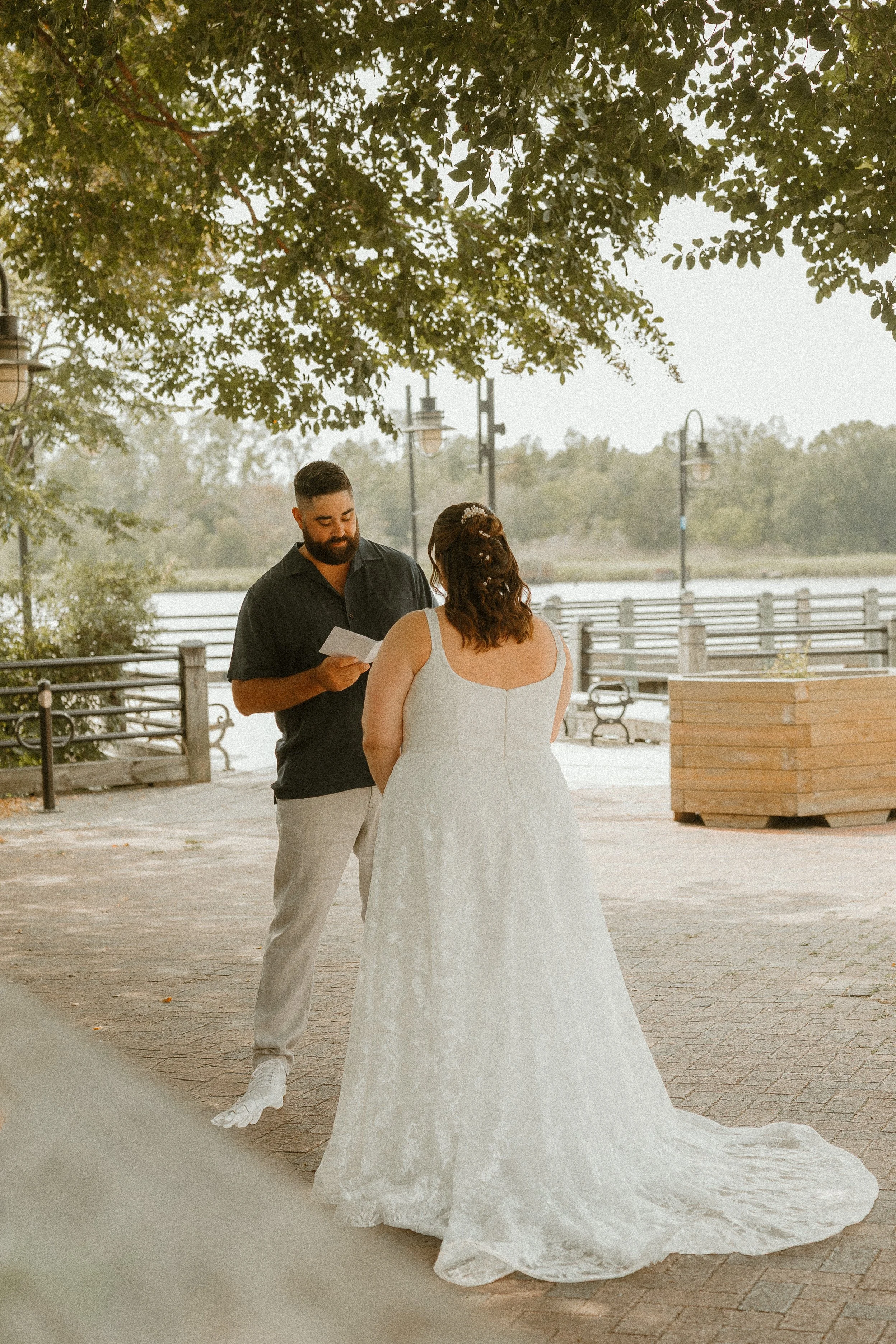 A man and woman dressed in wedding attire sharing a moment outdoors near a river, with the man reading a note and the woman in a white wedding gown.