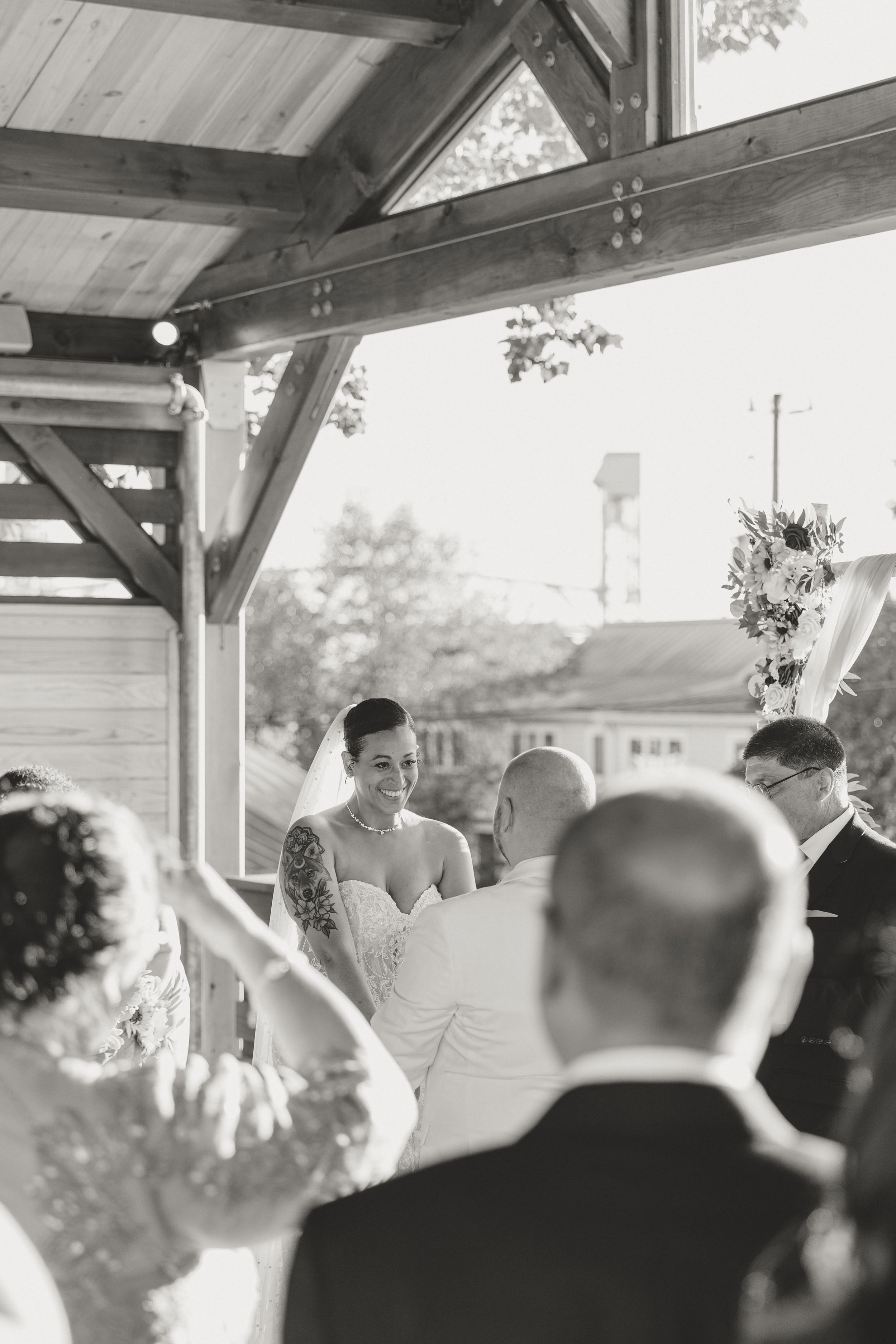 Black and white photo of a bride smiling at her wedding ceremony under a wooden structure outside, with guests seated around her.