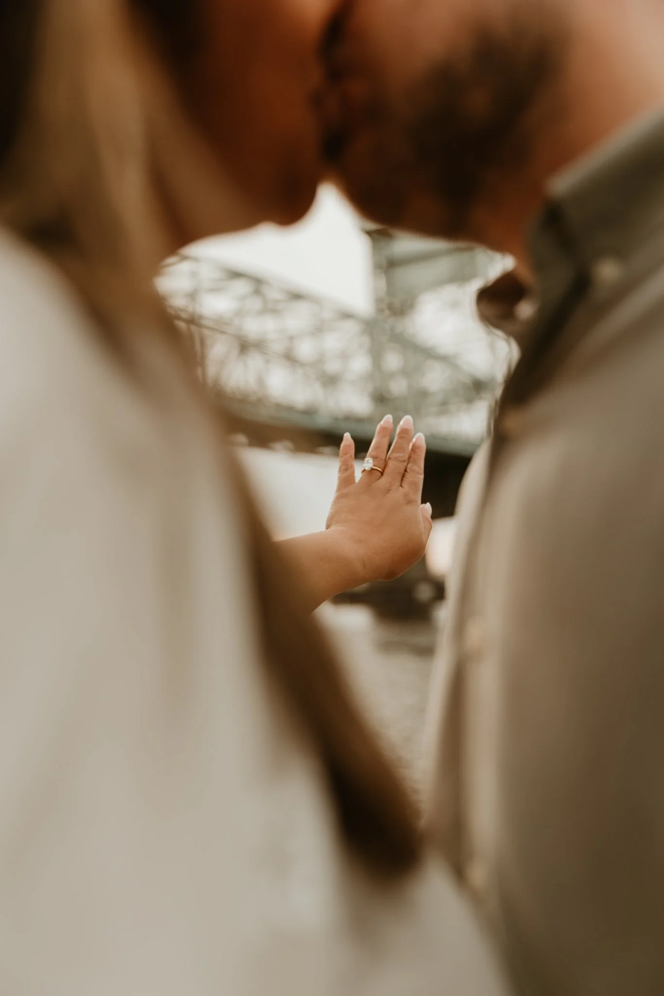Close-up of a couple kissing, with a focus on the woman's hand showing an engagement ring, in an outdoor setting.