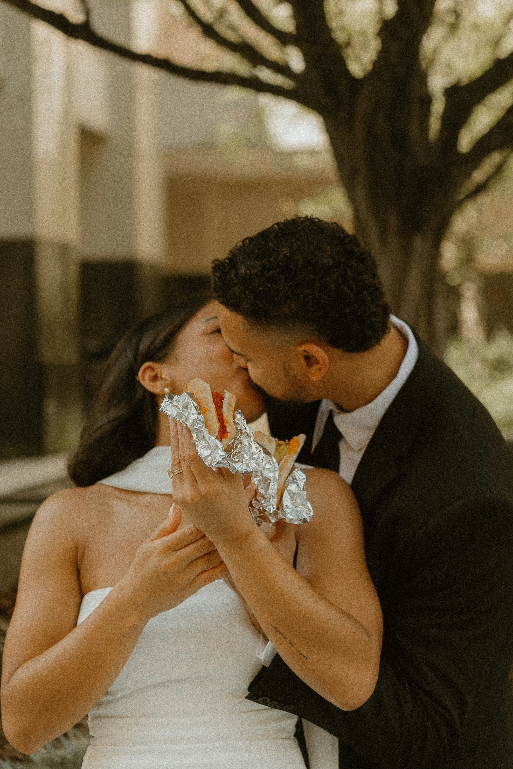 A couple sharing a kiss during a wedding ceremony, with the bride holding a bouquet wrapped in aluminum foil.