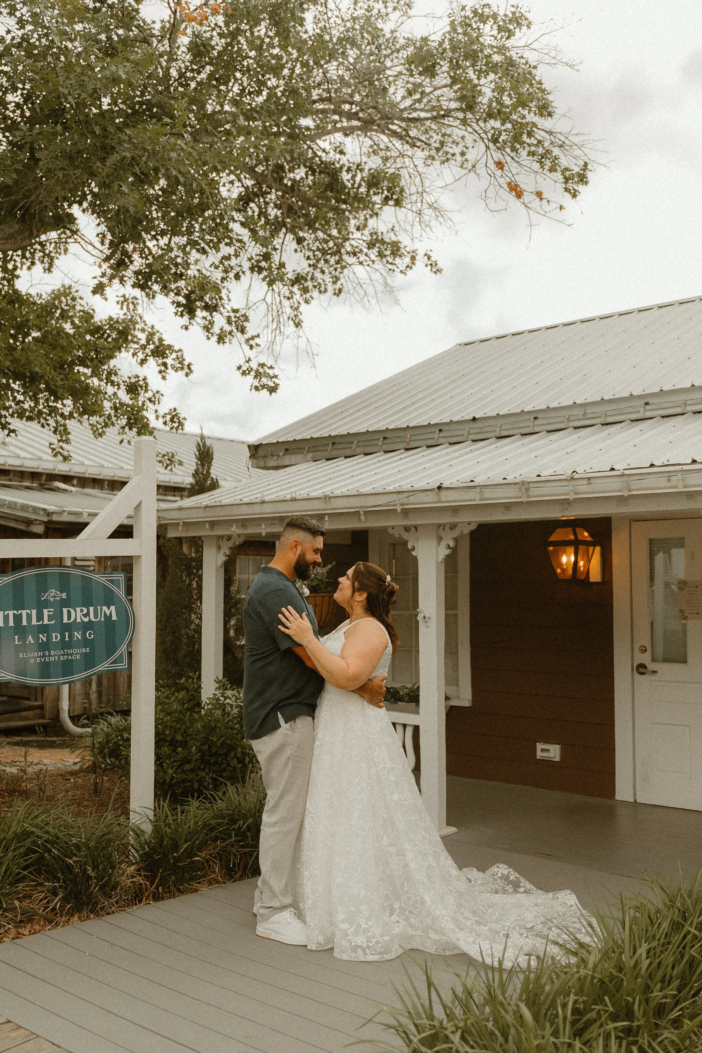 A bride and groom are standing close and holding each other while looking into each other's eyes outside a building with a sign that reads 'Little Drum Landing.' The bride is wearing a white lace wedding gown with a train, and the groom is dressed in