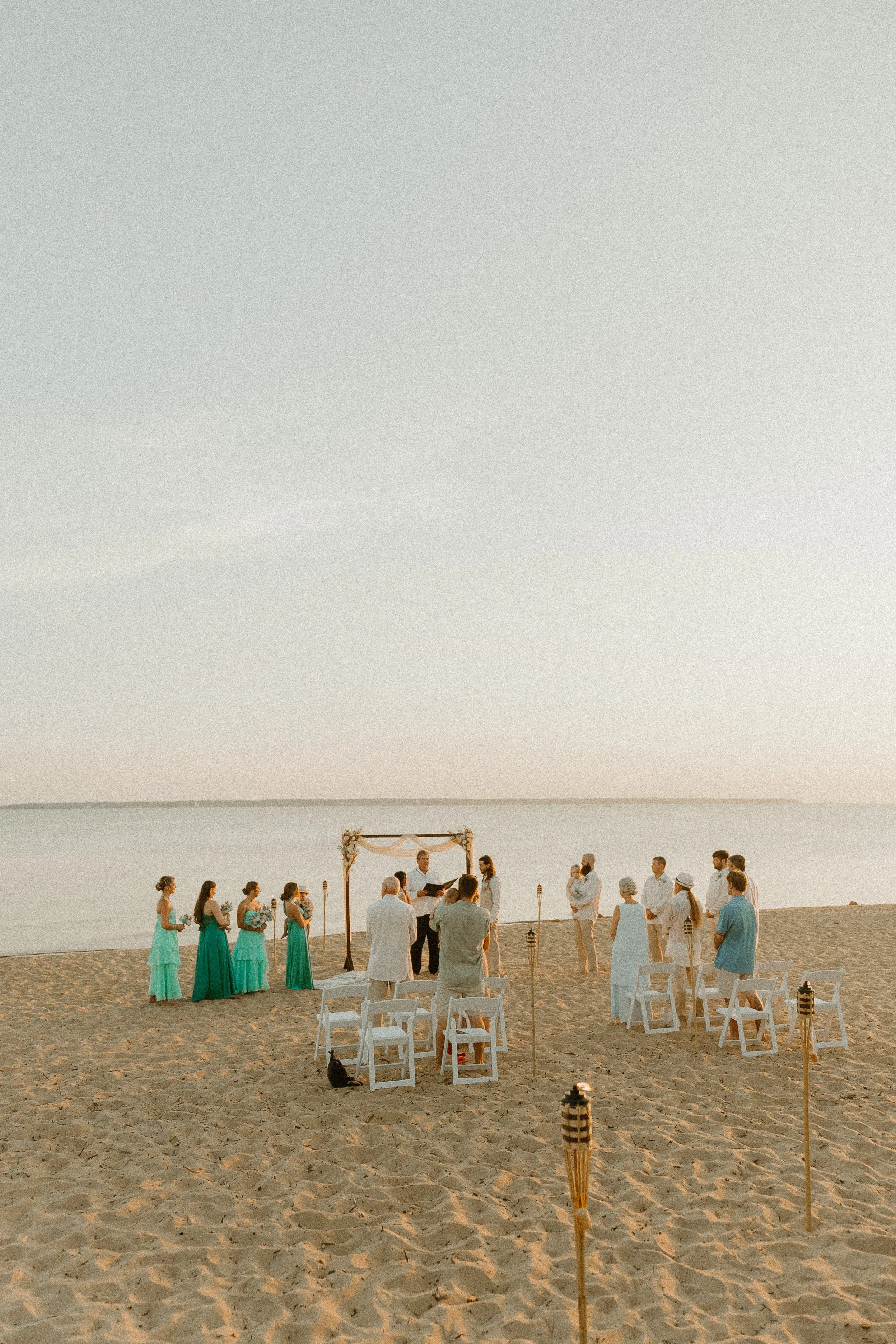 A beach wedding ceremony with a group of people gathered around an arch, by the water at sunset, with chairs, tiki torches, and guests in formal attire.