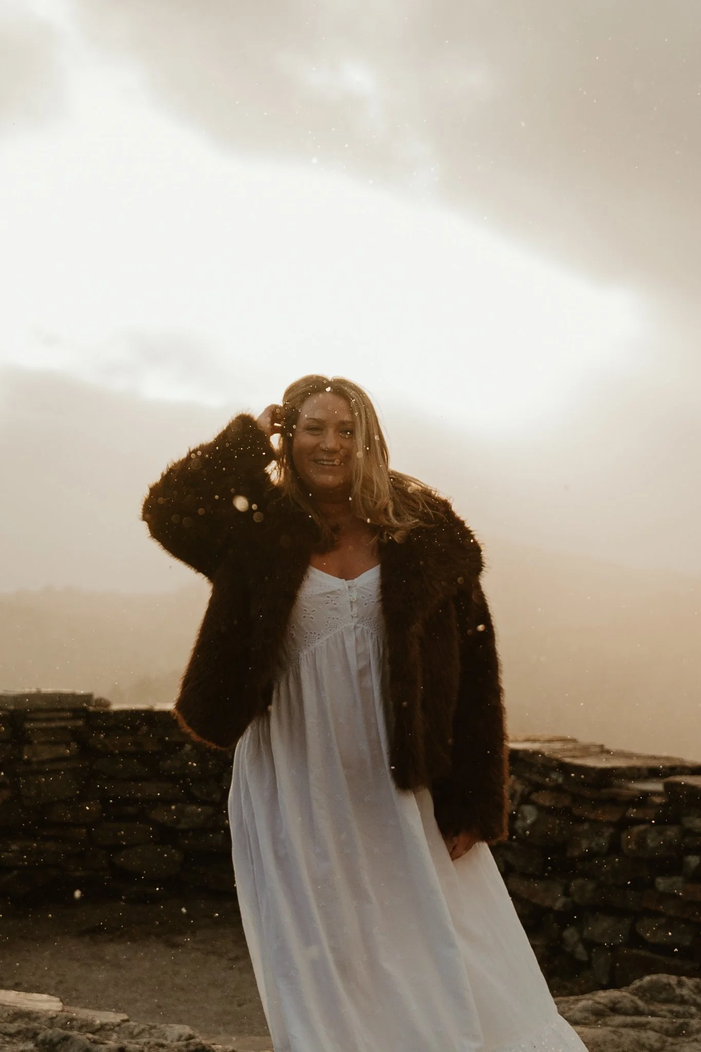 A woman wearing a white dress and a dark fur coat standing outdoors near a stone wall during rainy weather, with raindrops visible in the air and a cloudy sky in the background.