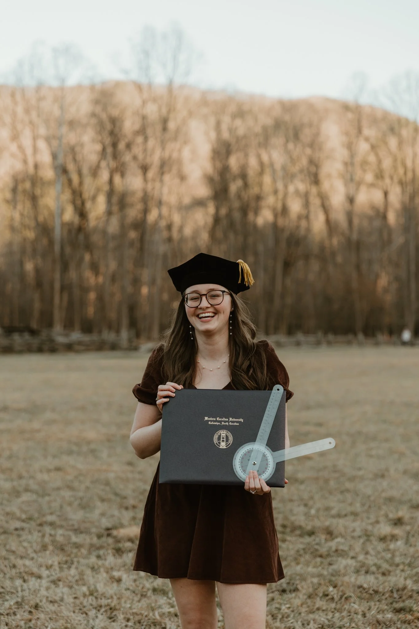 A young woman in graduation cap and gown holding a black diploma cover and white directional compass outdoors on a grassy field with trees in the background.