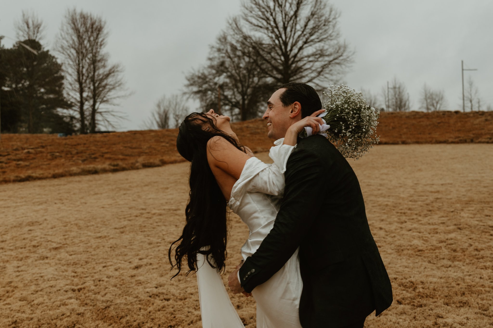 A couple in wedding attire sharing a joyful moment outdoors on a cloudy day, with the woman holding a bouquet of baby's breath flowers.