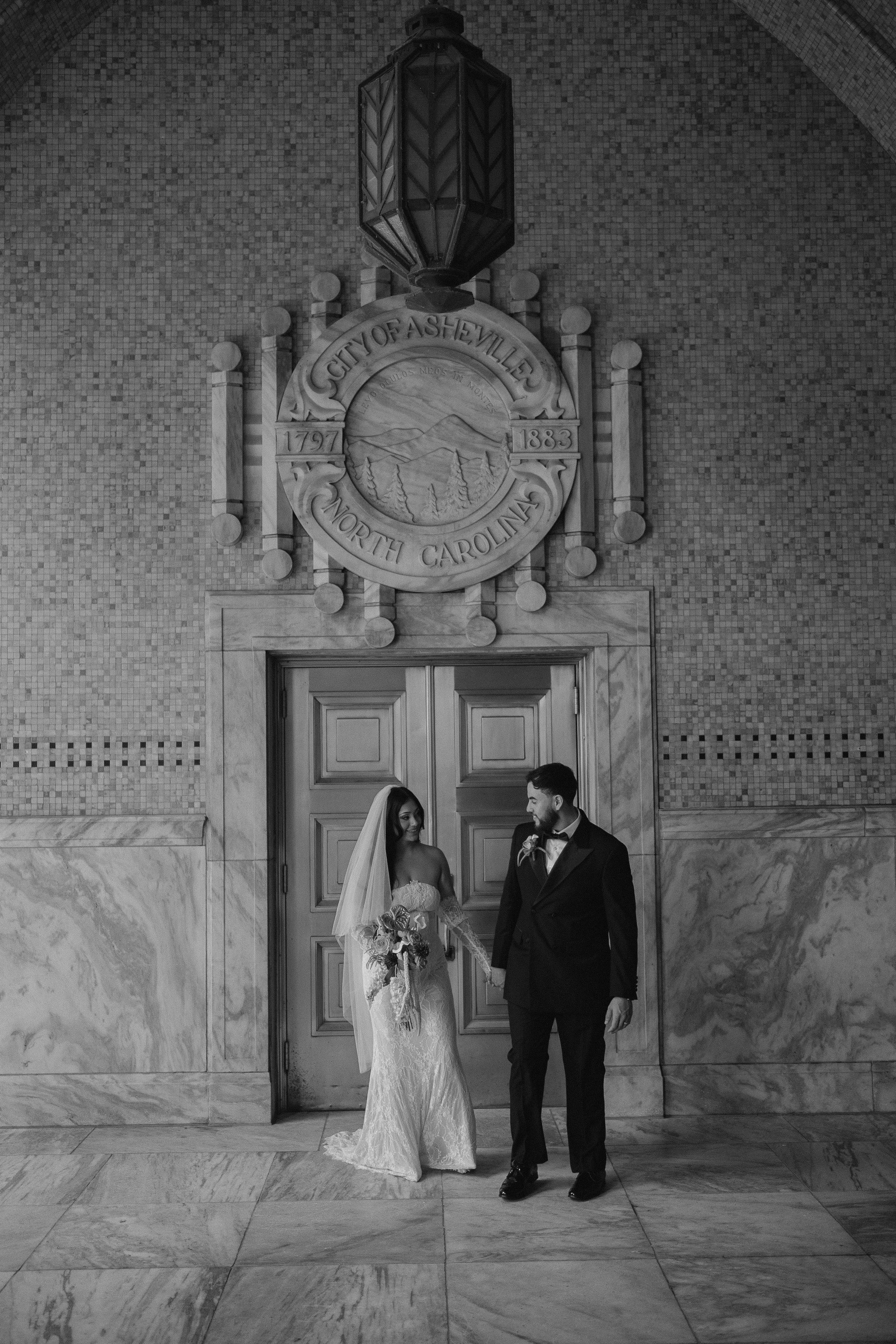 A bride and groom standing and holding hands in front of a wooden door inside a building with a tile wall and a sign above them that reads 'City of Asheville North Carolina' with the years 1791 and 1883. The bride is wearing a lace wedding dress and veil, holding a bouquet, and the groom is wearing a dark suit, white shirt, and tie.