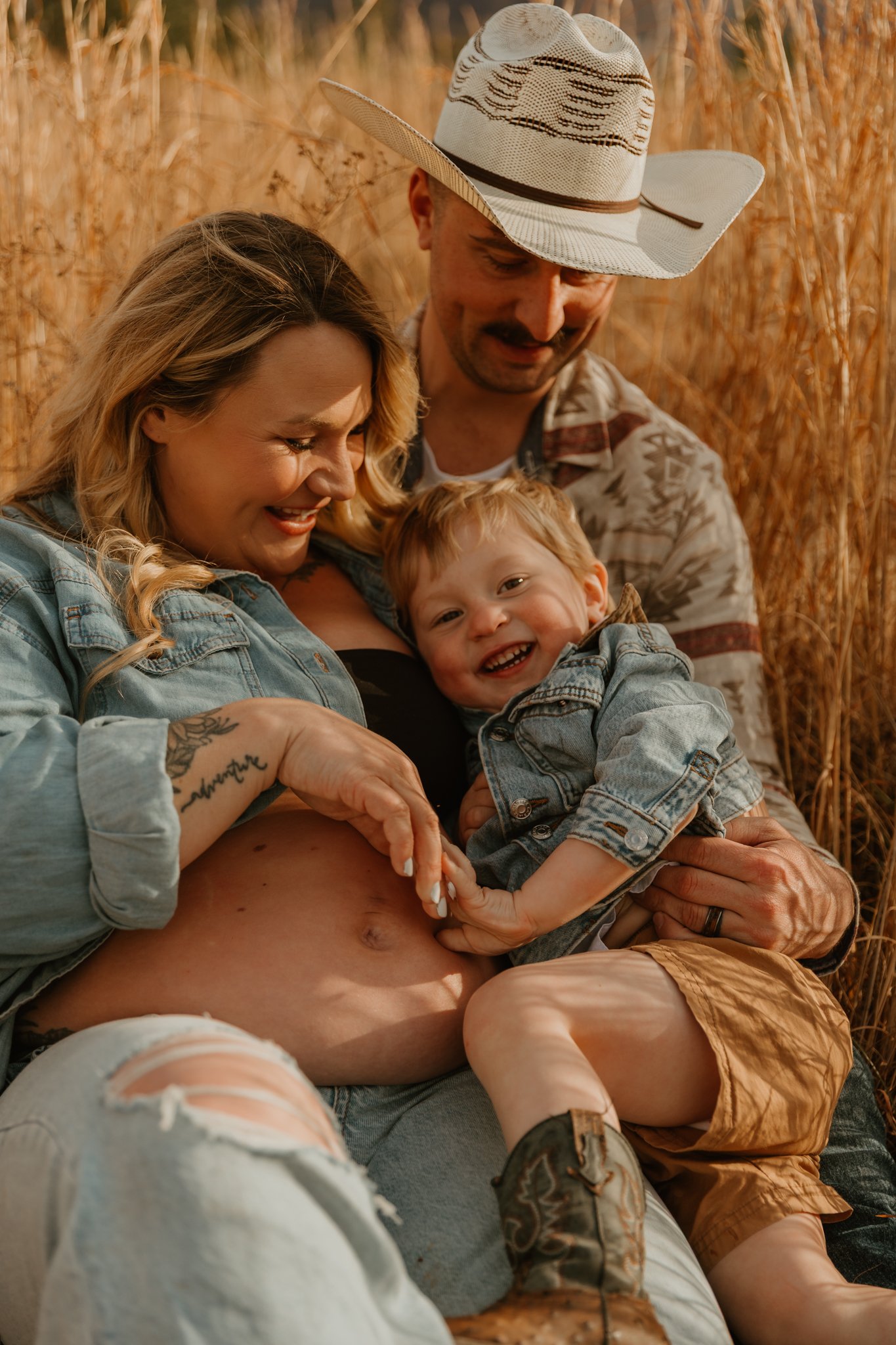 A smiling family of three, consisting of a pregnant woman, a man, and a young boy, sitting in a field of tall, golden grass. The woman is touching her belly while the boy is playfully touching her stomach, and they are all enjoying a joyful moment to