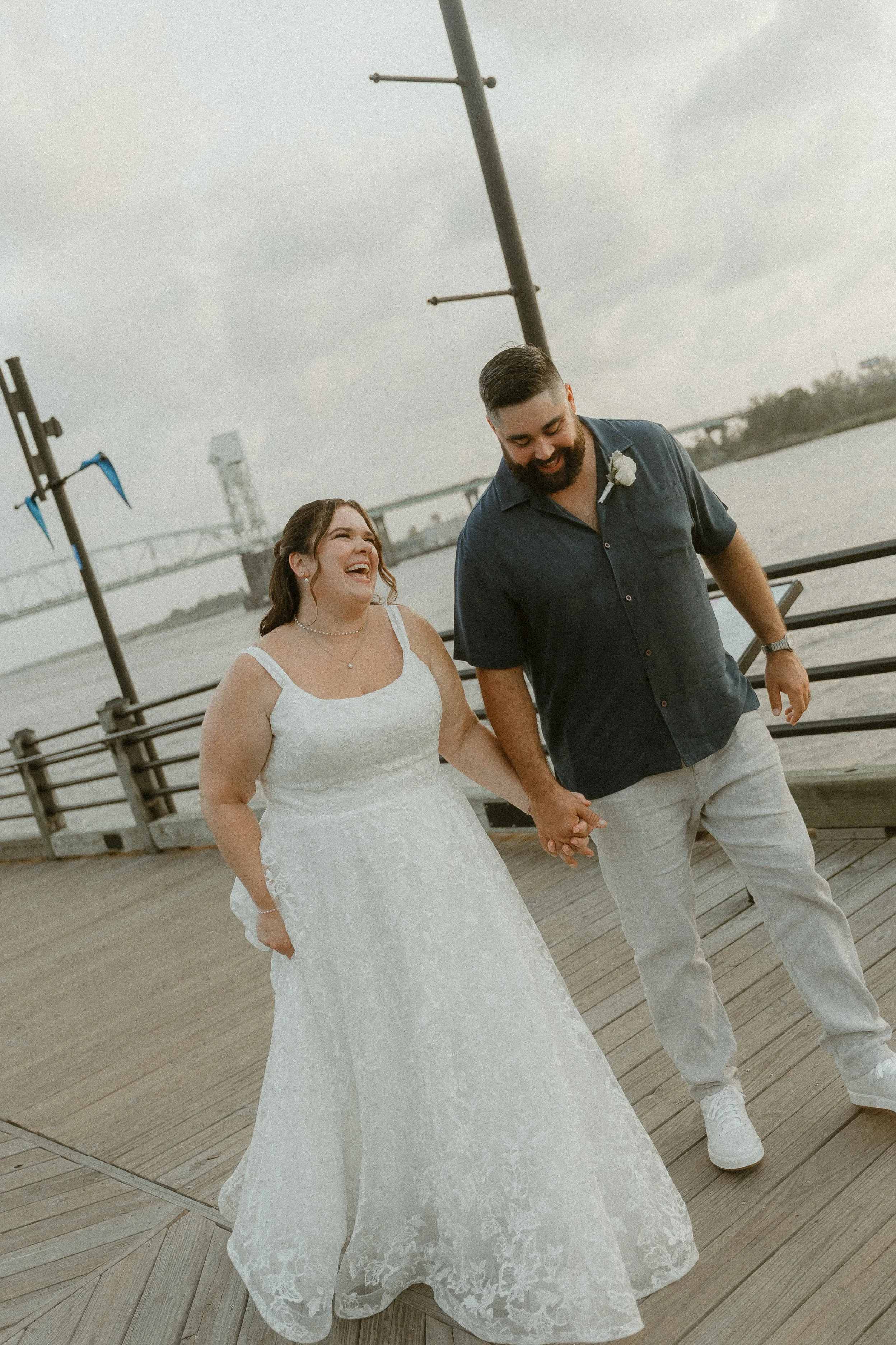A smiling bride and groom holding hands on a wooden pier near the water, with a bridge and cloudy sky in the background.