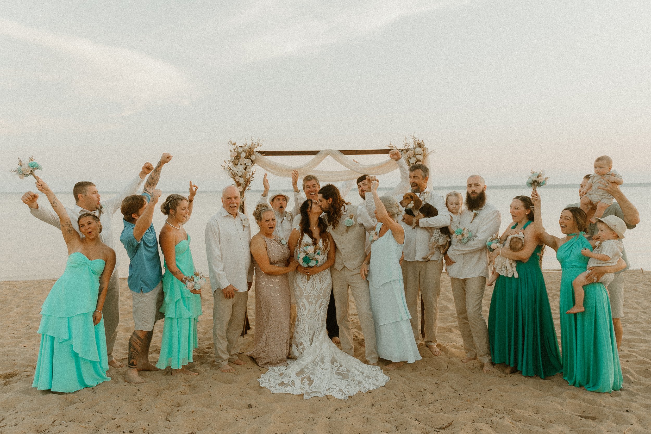 A group of people on a beach celebrating a wedding, with the bride and groom at the center under a decorated arch, surrounded by friends and family, some with children, all dressed in summer attire.