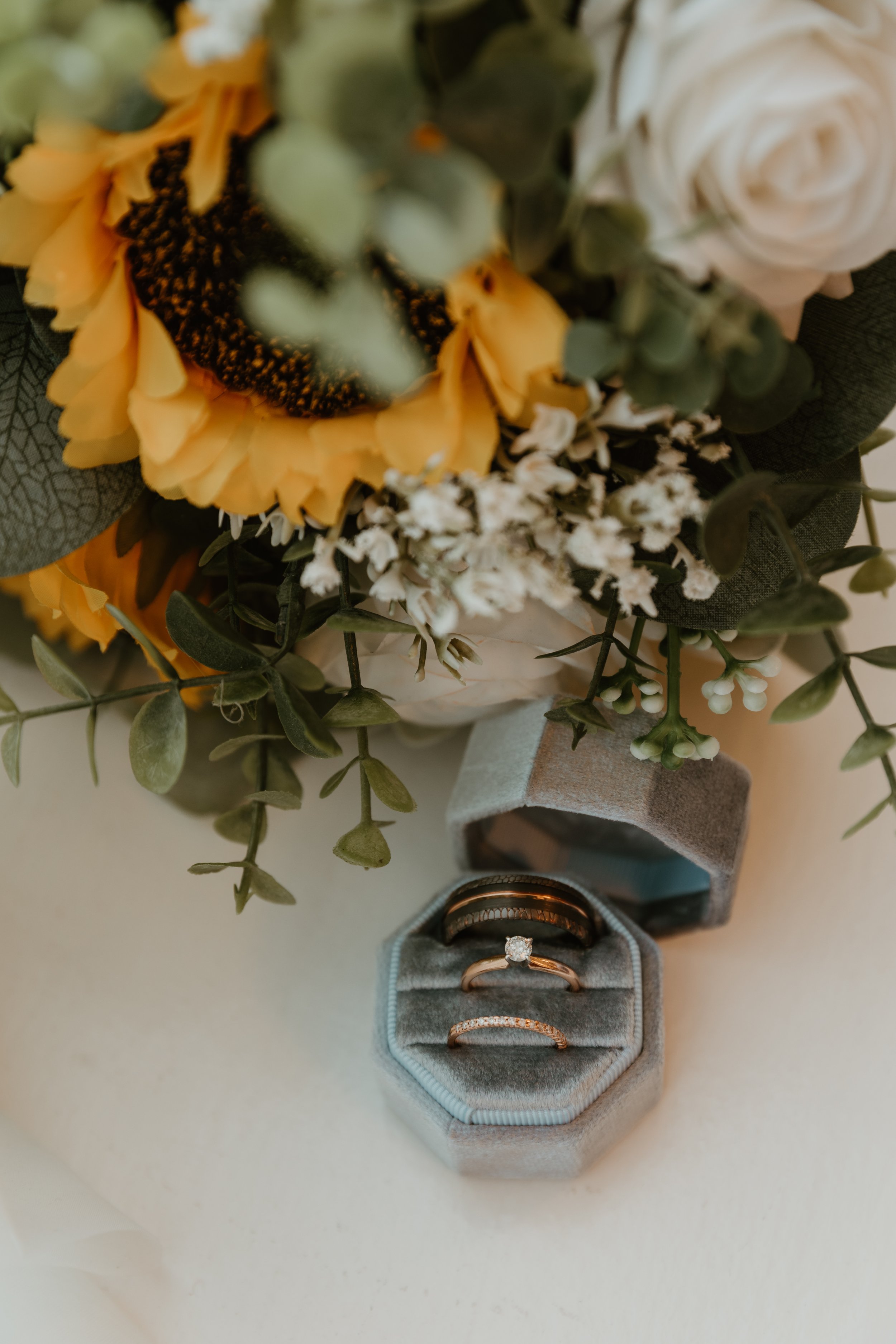 A bouquet of flowers with sunflowers, white roses, and greenery placed next to a gray jewelry box containing a diamond engagement ring and wedding band.