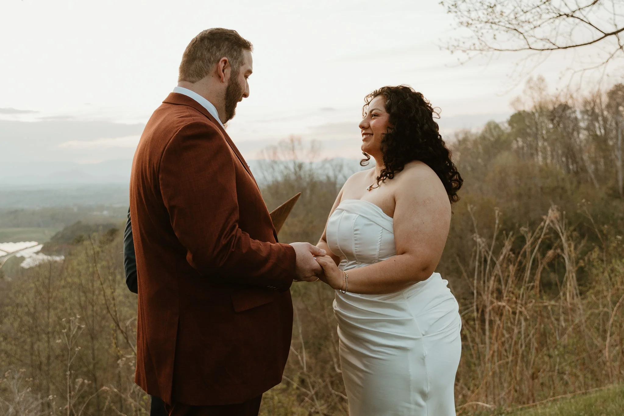 A man and woman share an emotional moment outdoors, holding hands, with the man wearing a brown jacket and the woman in a white dress, against a backdrop of trees and a cloudy sky.