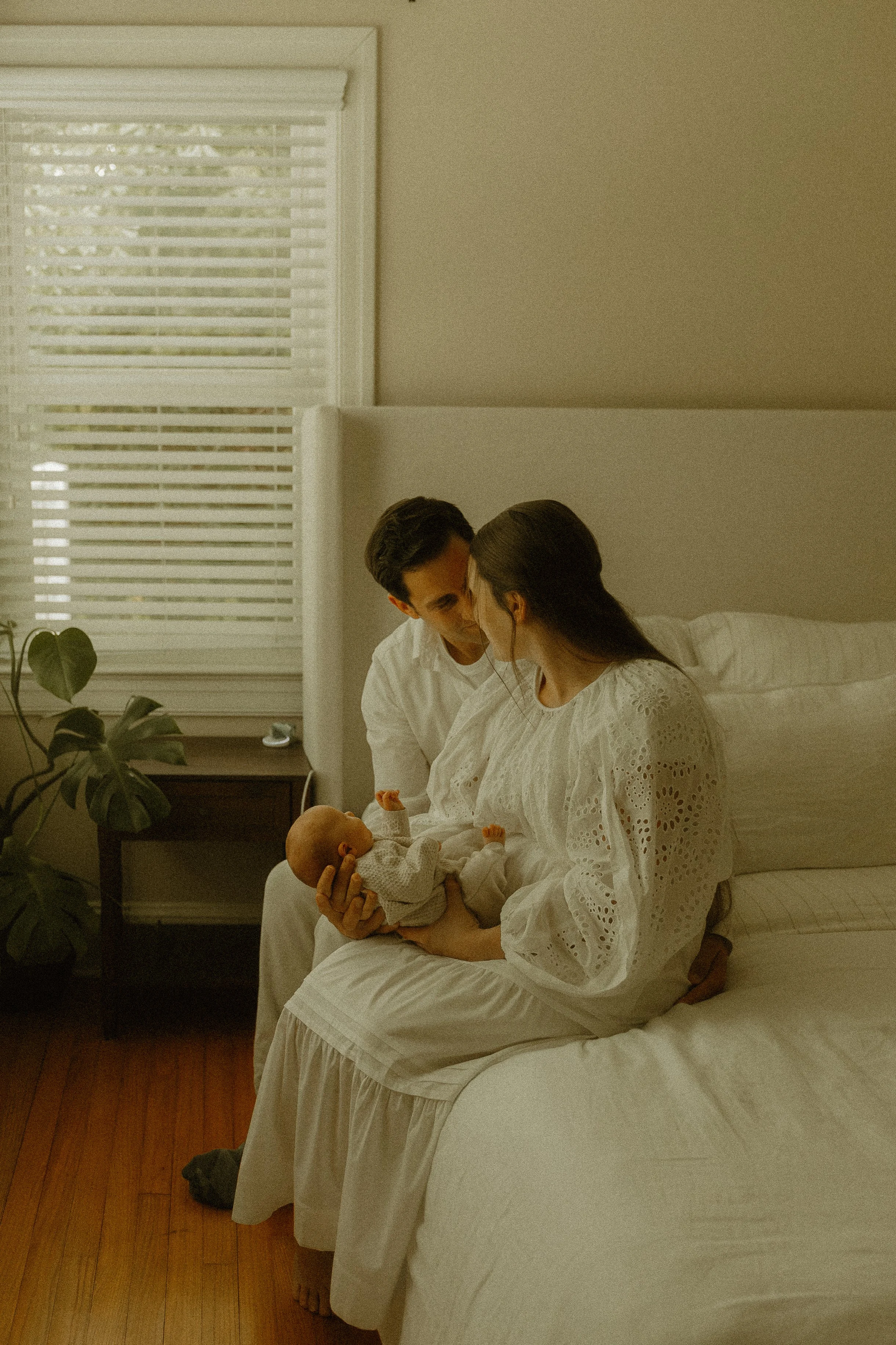 A couple sitting on a bed holding a baby doll in a room with white walls, a window with blinds, and a plant on a nightstand.