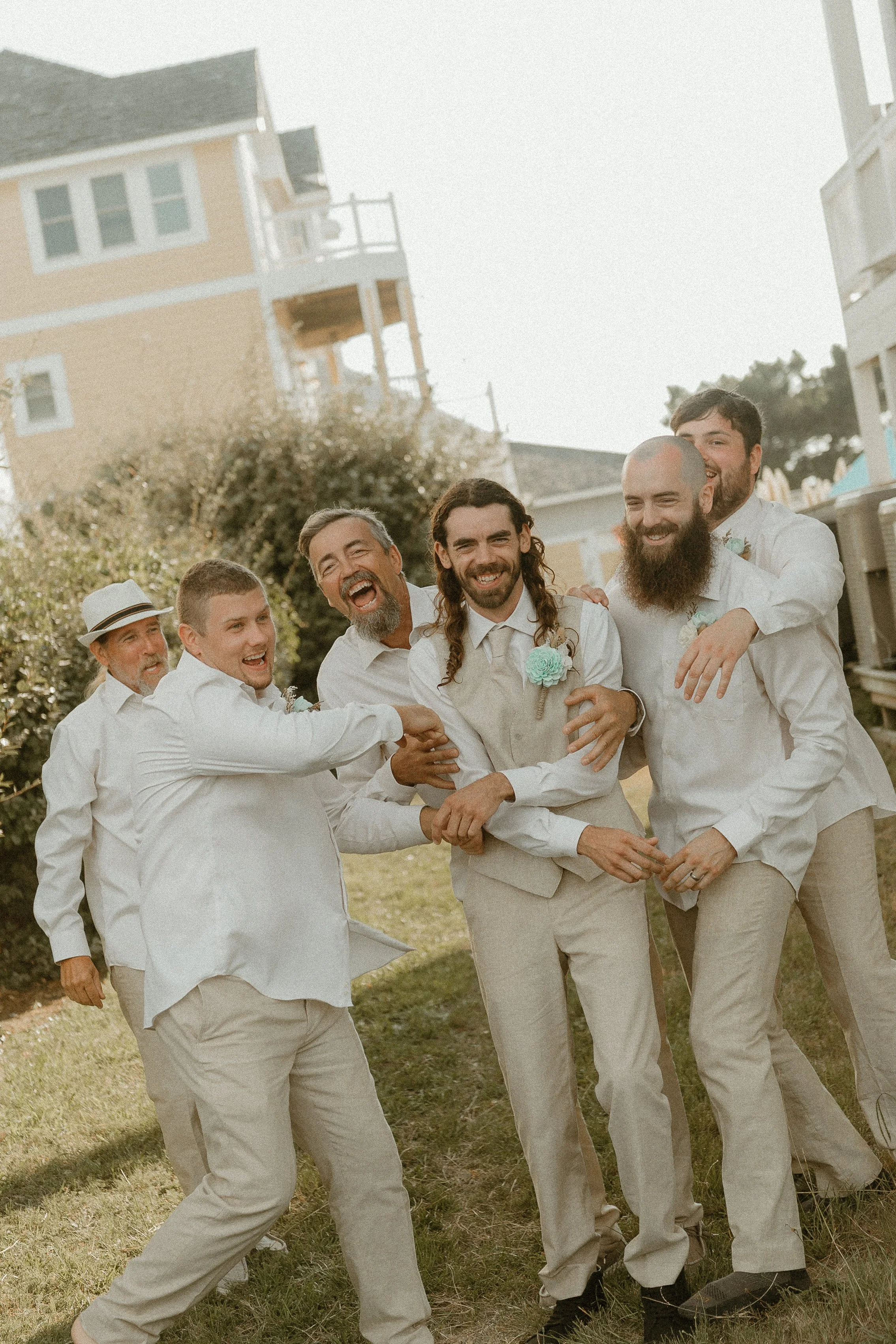 Group of men dressed in light-colored suits and shirts, smiling and laughing outdoors during daytime, some with floral boutonnières, with houses and trees in the background.