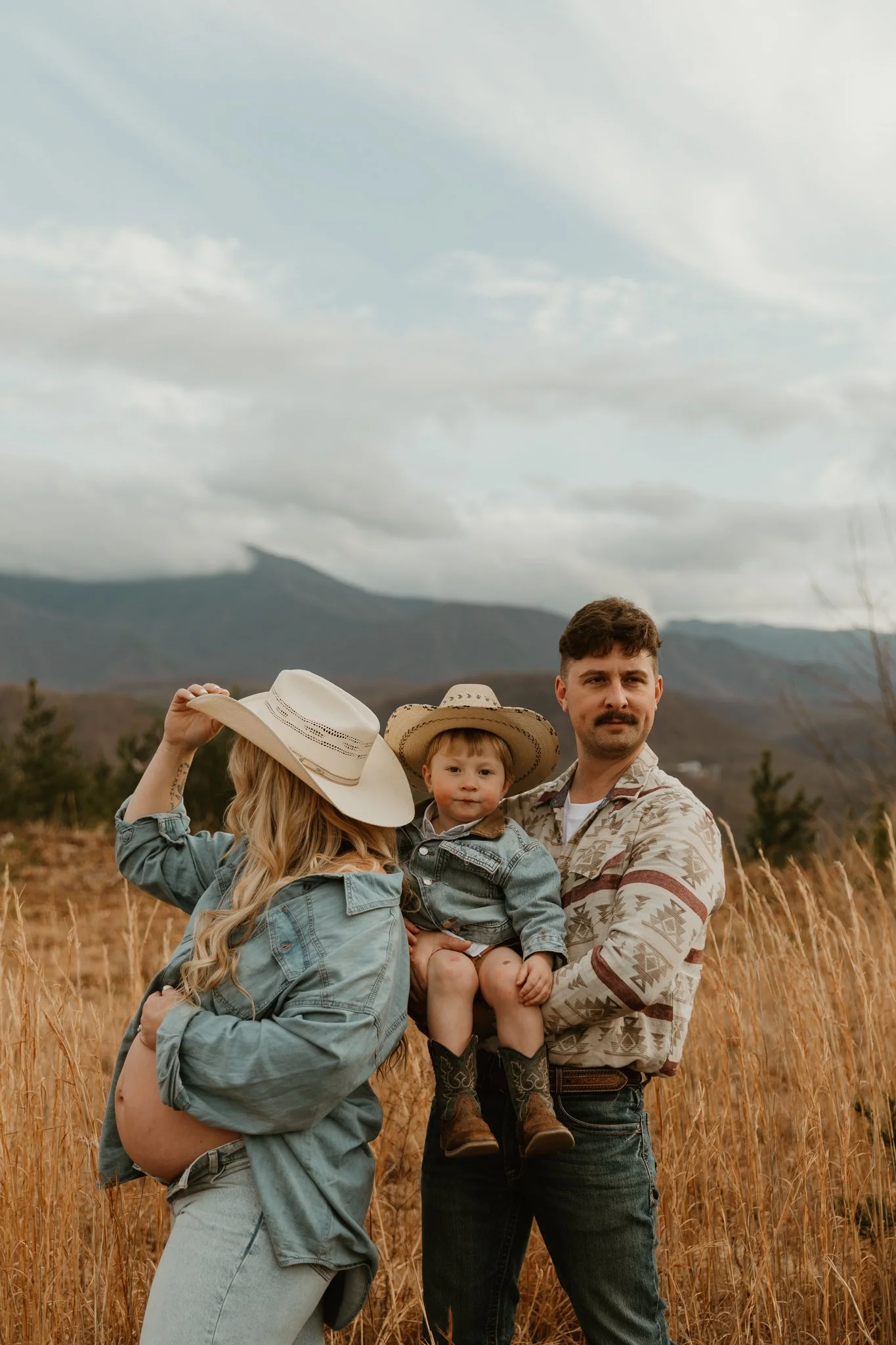 A family of three outdoors in a field of golden grass with mountains in the background. The woman is holding a small child, with the woman wearing a large hat and the man holding the child. The woman is wearing a cowboy hat and denim jacket, while th