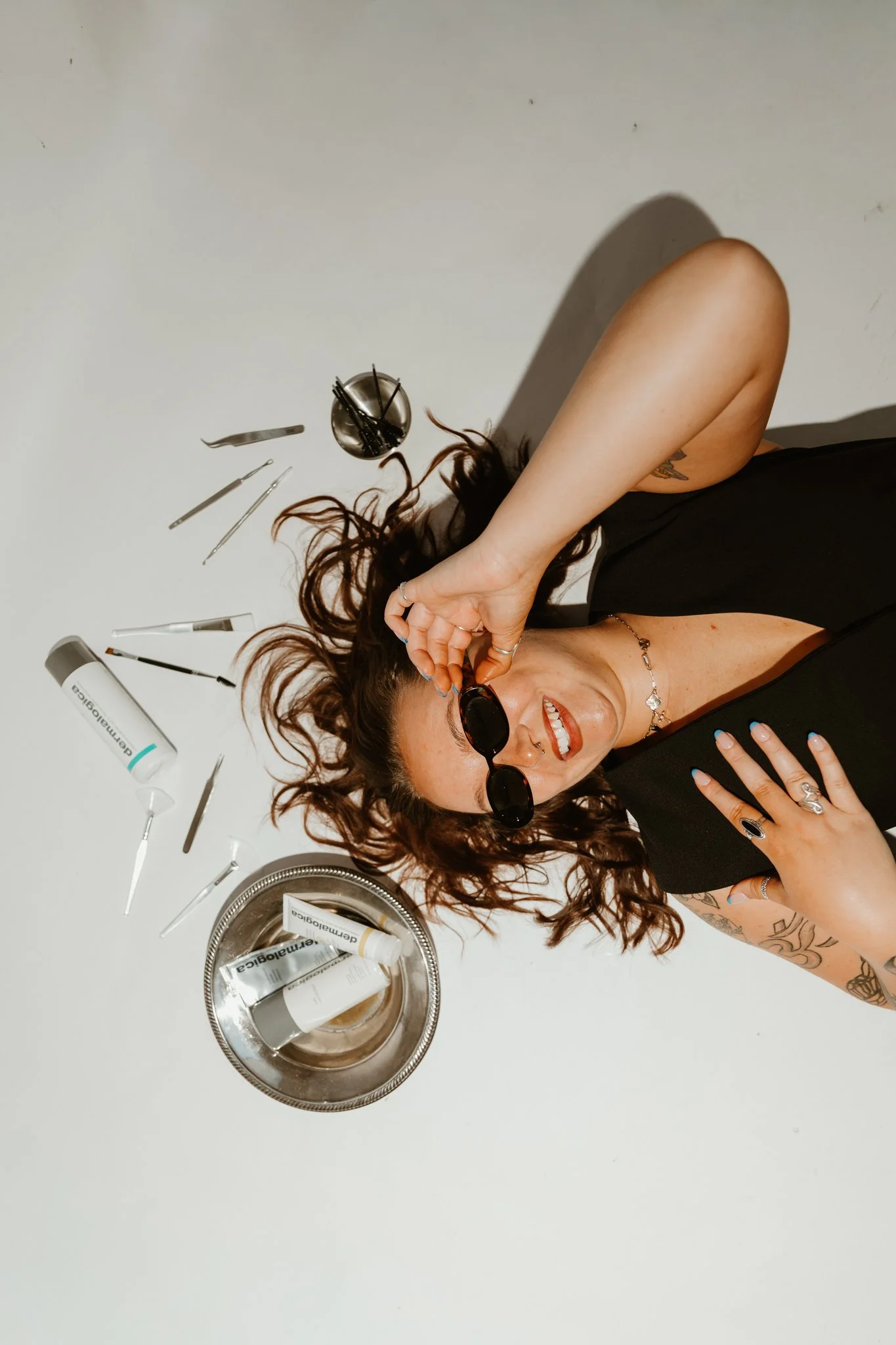 A woman lying on a white surface, wearing sunglasses, surrounded by tattooed arms, with various skincare and beauty tools and products nearby.