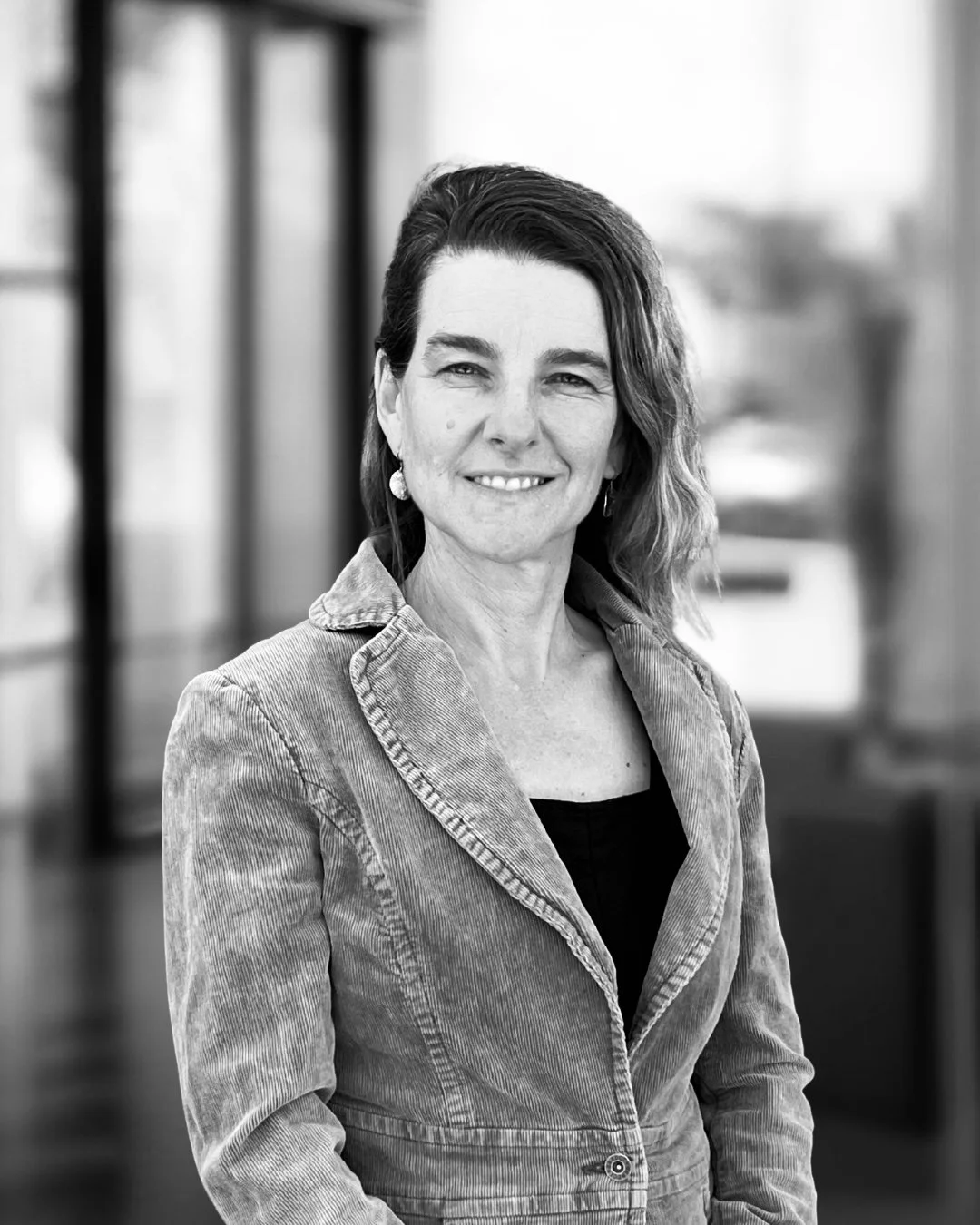 Black and white portrait of a woman smiling, wearing glasses, earrings, and a patterned top, sitting indoors with windows in the background.