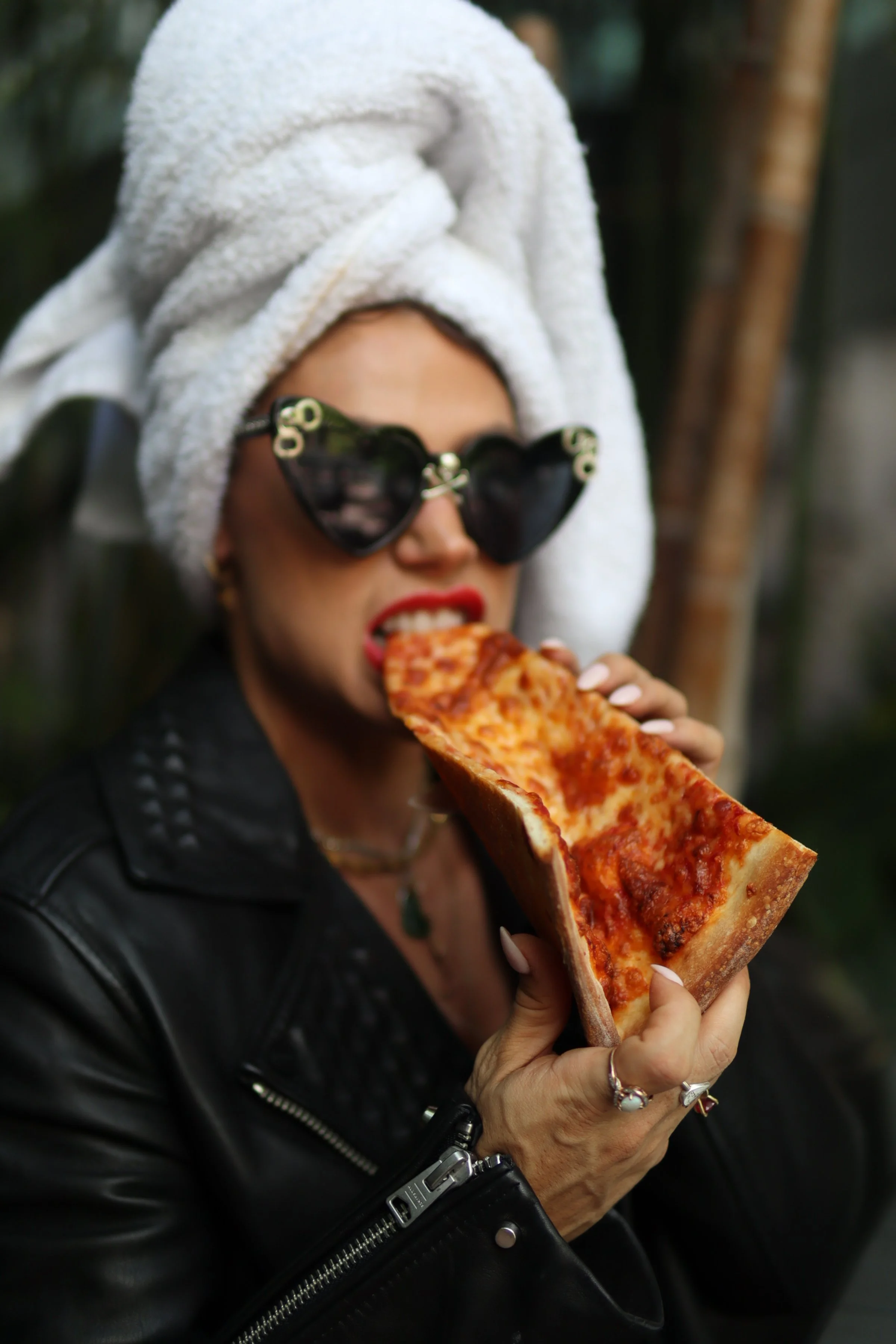 Woman with a white towel wrapped around her head, wearing heart-shaped sunglasses, a black leather jacket, and jewelry, holding a slice of pizza with pepperoni and cheese close to her mouth.