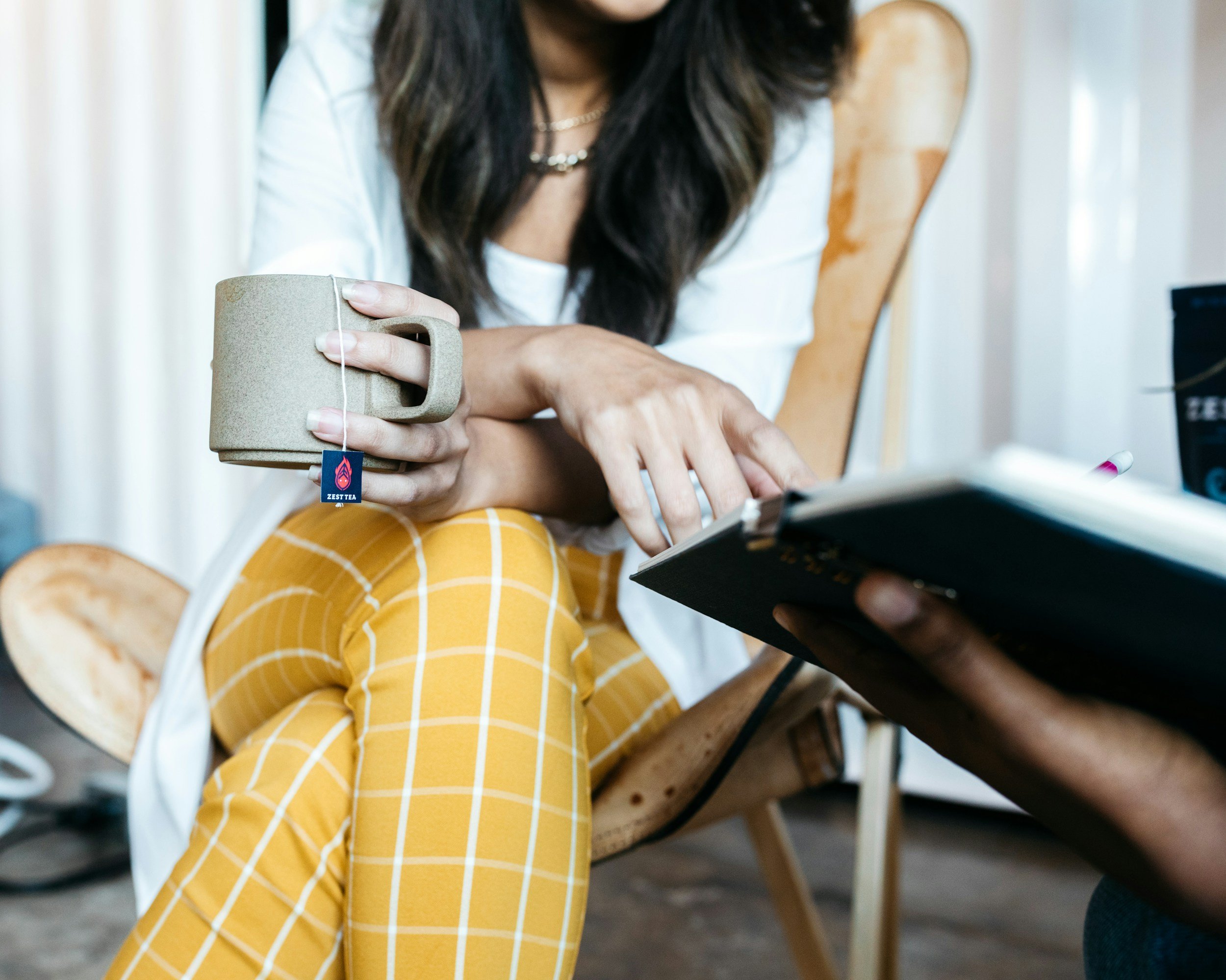 A woman with long dark hair, yellow plaid pants, and a white shirt is sitting on a wooden chair, holding a mug labeled 'Zest Tea' in her right hand and writing or reading in a notebook with her left hand.
