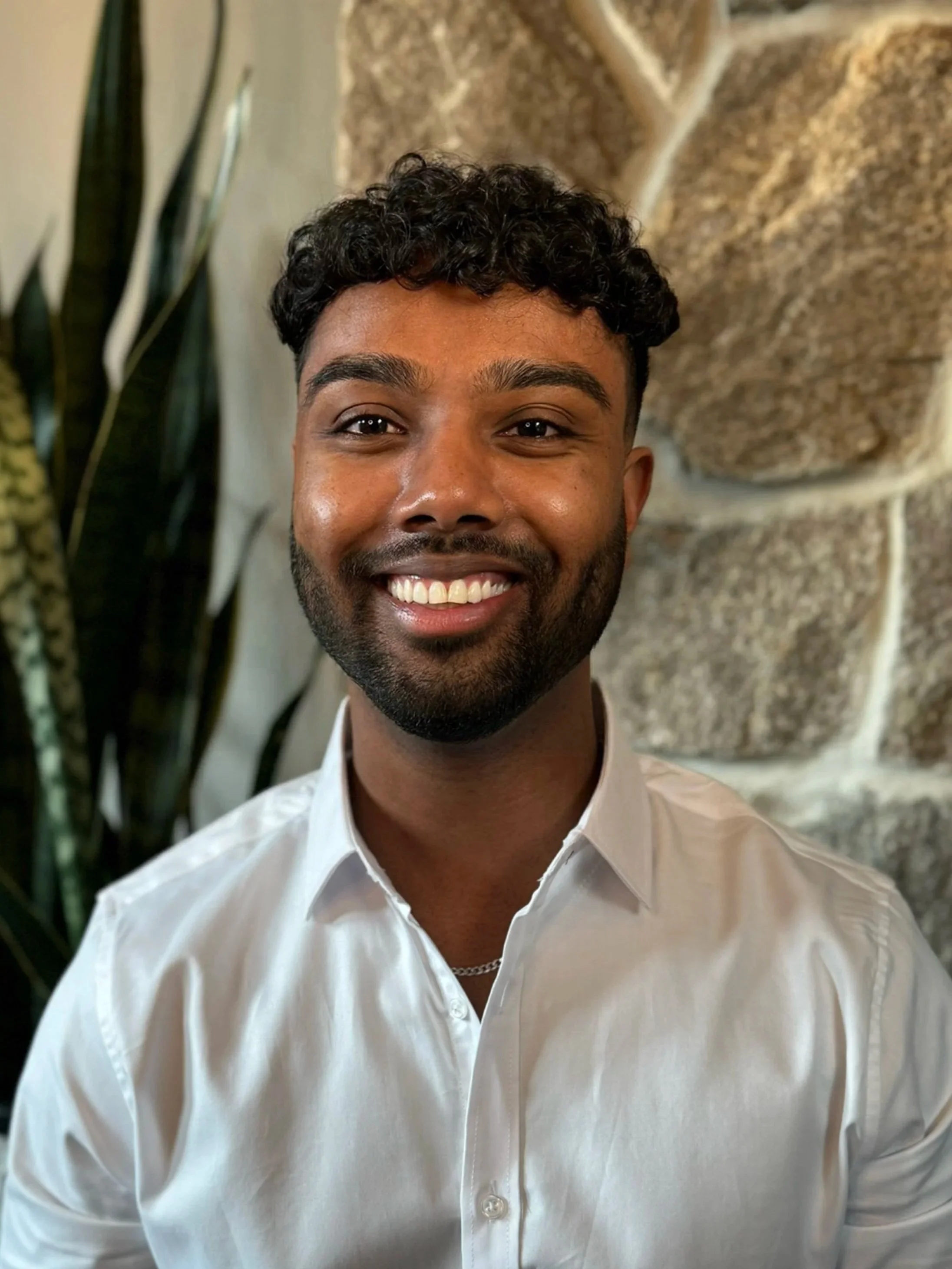 A smiling young man with curly black hair and a beard, wearing a white dress shirt, standing in front of a stone wall and a plant.