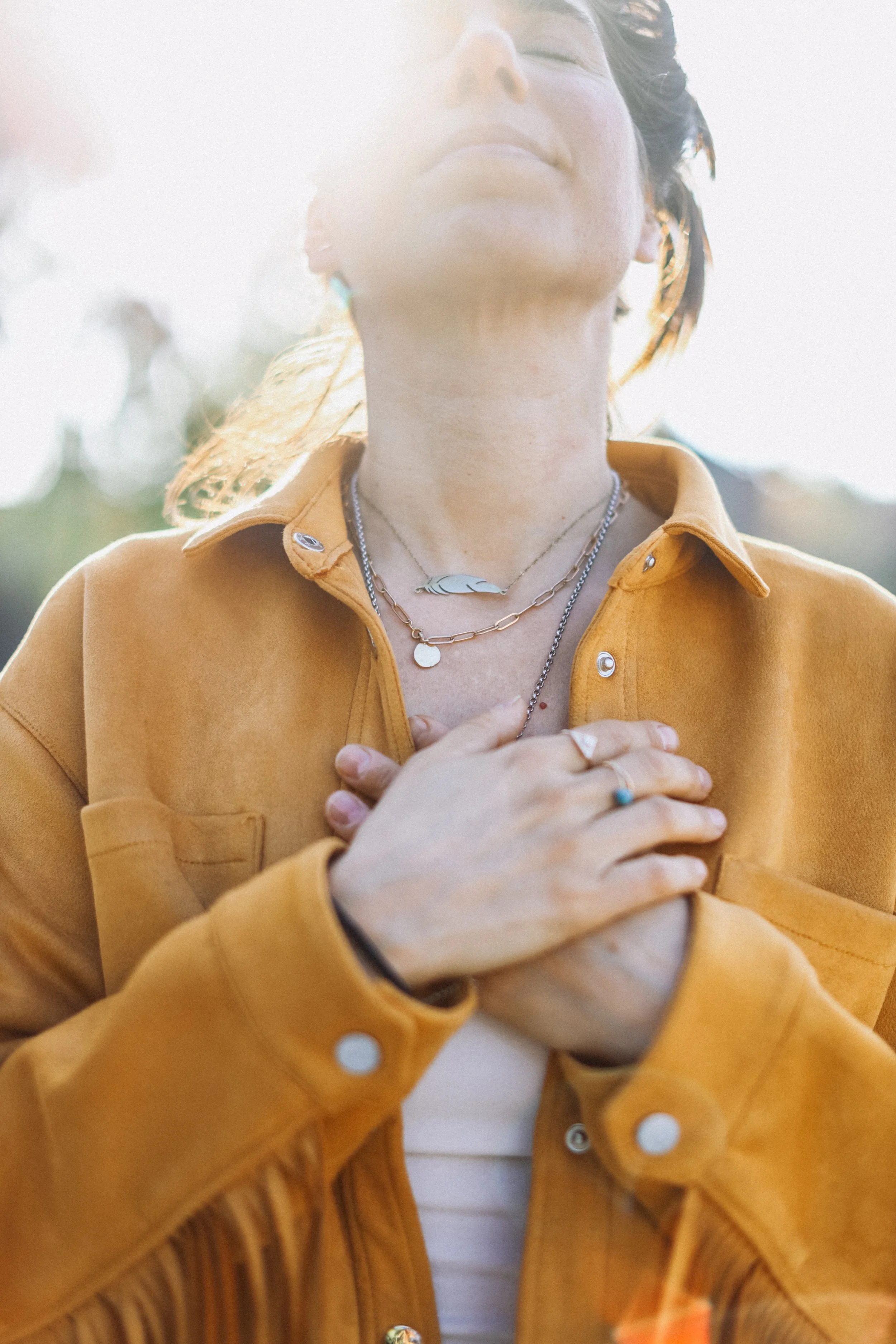 A woman outdoors with sunlight shining on her, wearing a mustard-yellow jacket and jewelry, with her hands crossed over her chest.