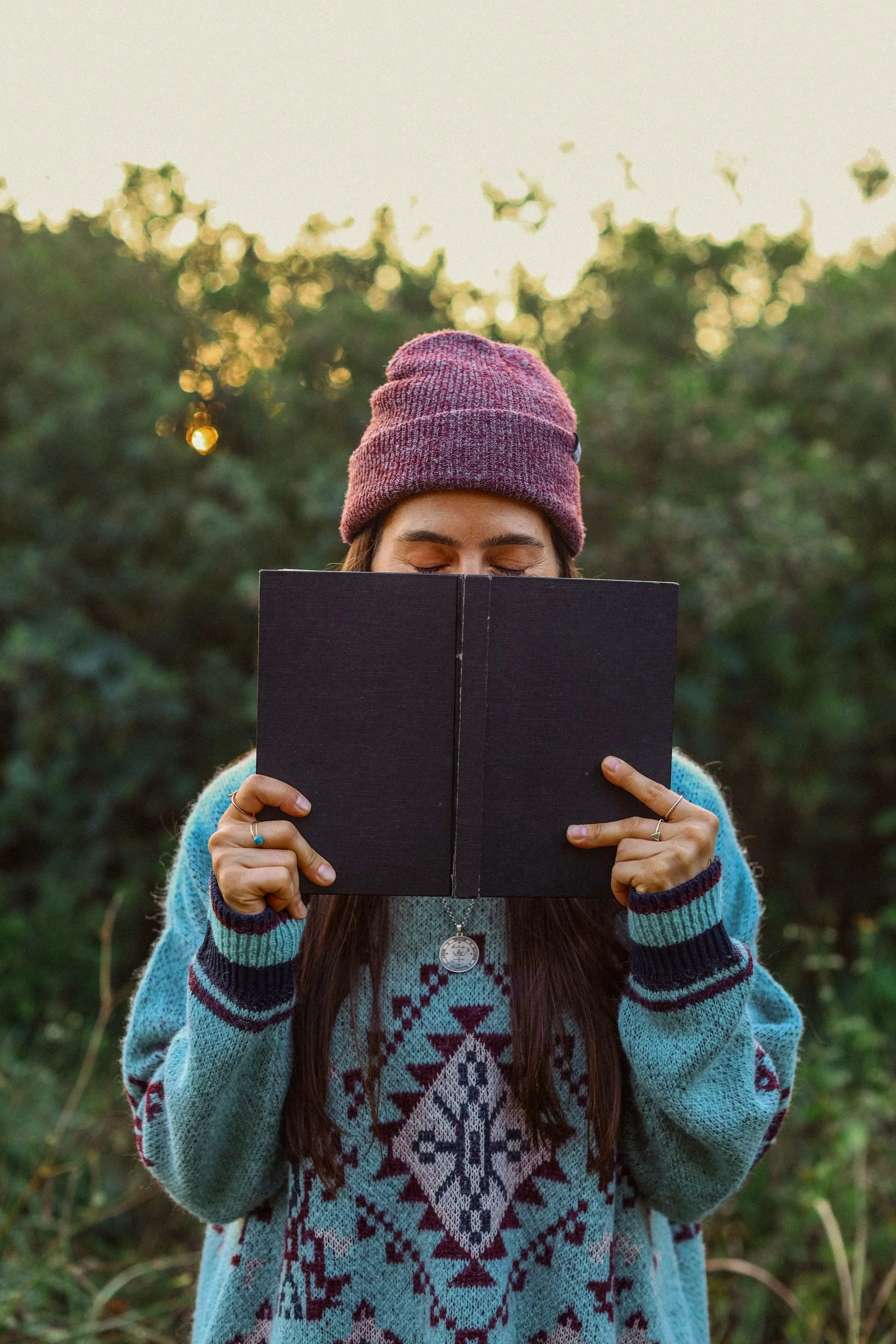 Woman in a colorful sweater and purple beanie reading a black notebook outdoors during sunset.