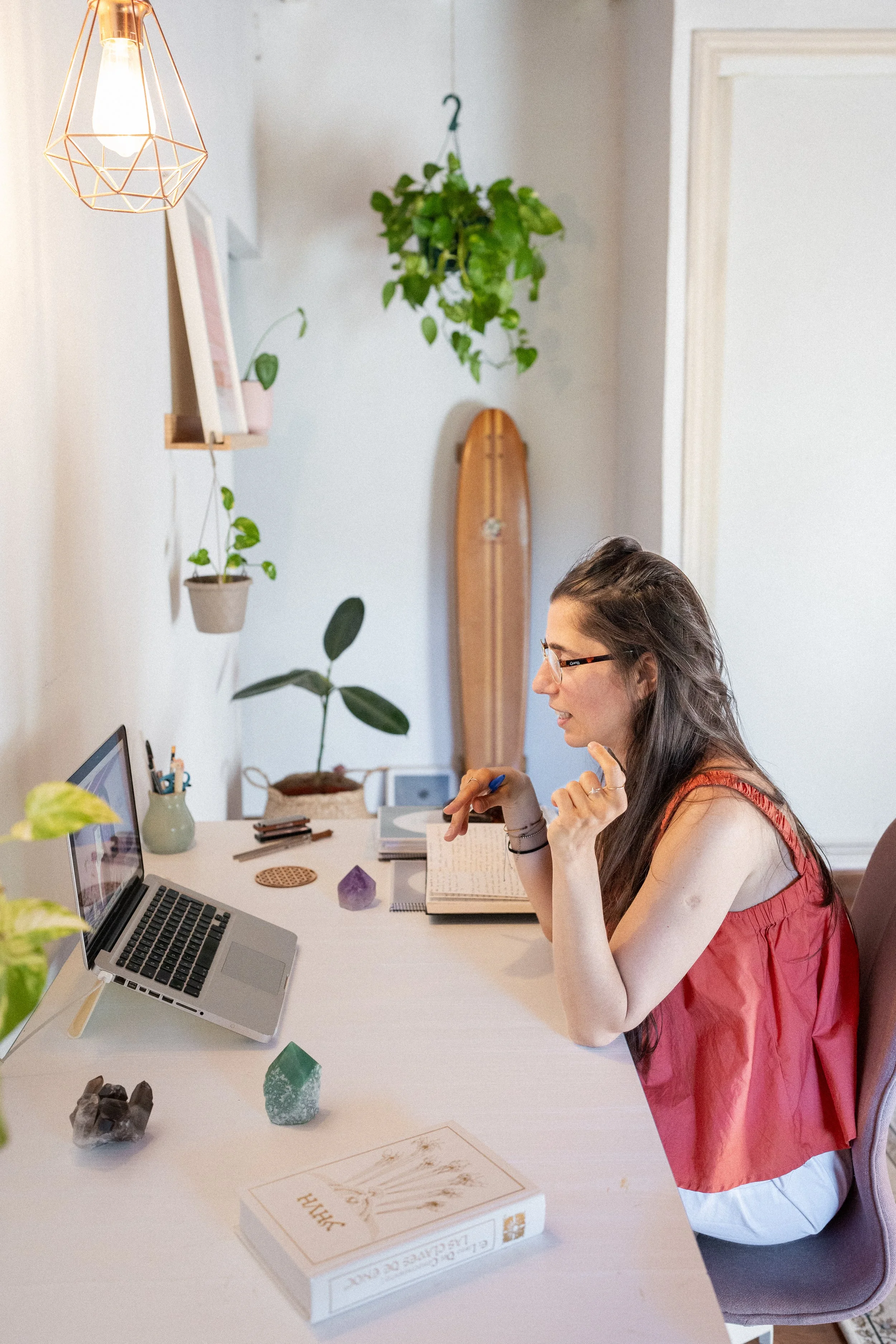 A woman with long dark hair and glasses, wearing a sleeveless red top, sitting at a desk working on a laptop. The desk has plants, books, crystals, and stationery. A hanging lamp and wall decor are visible in a brightly lit, cozy room.