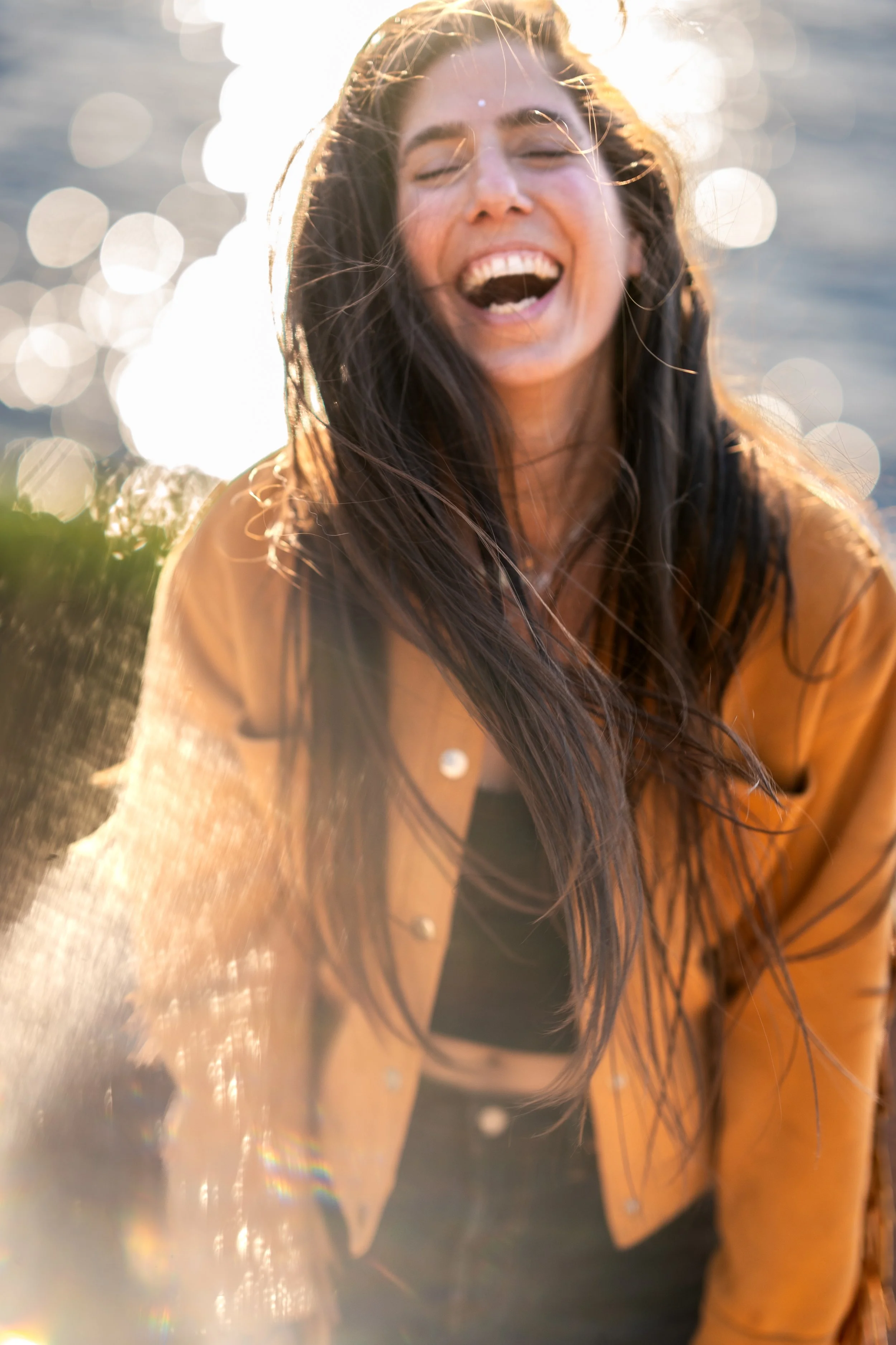 A woman with long brown hair is outdoors near water, smiling and laughing, with sunlight reflecting off the water behind her.
