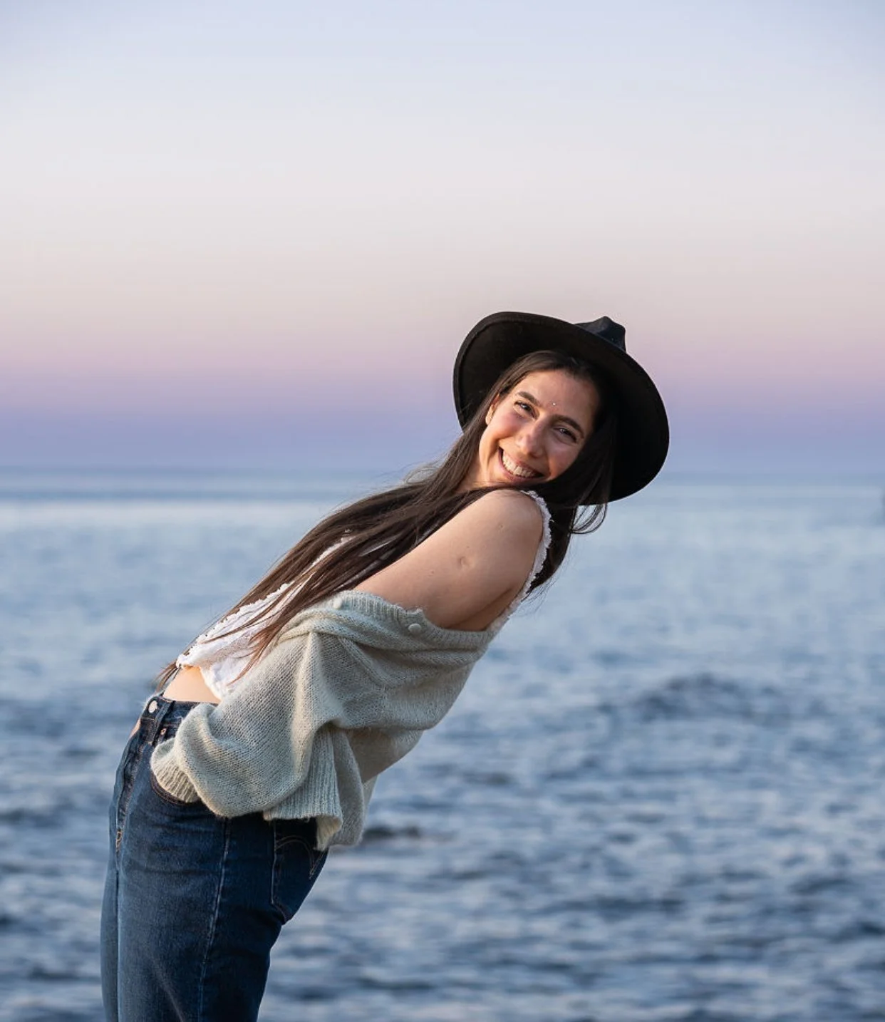 A woman with long hair wearing a black hat, smiling while leaning back near a body of water during sunset or dusk.