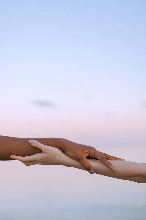 Two hands, one dark-skinned and one light-skinned, gently touching or holding each other's forearms against a soft pastel-colored sky background.