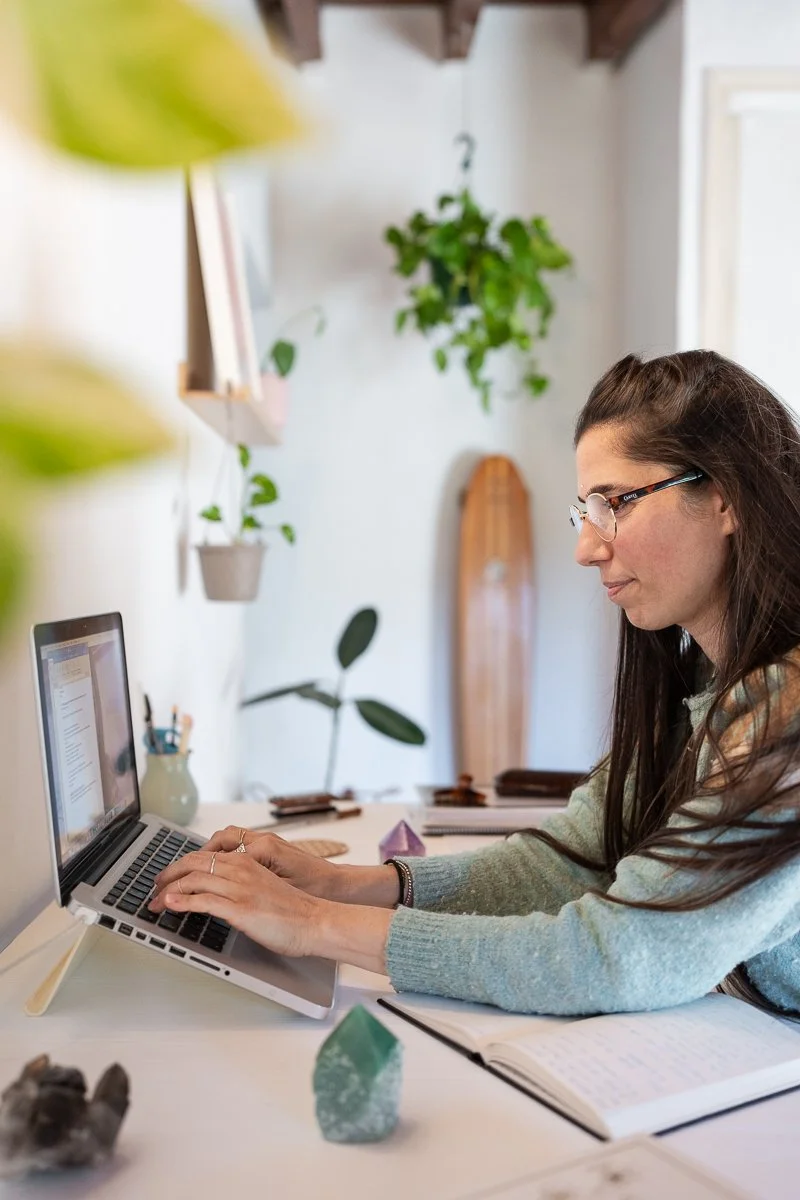 Woman working on a laptop at a desk with plants and notebooks in a cozy room