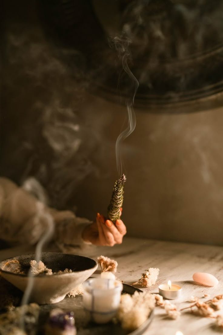 Close-up of a hand holding a smoldering incense stick with smoke rising, surrounded by candles and flower petals on a table in a dimly lit setting.