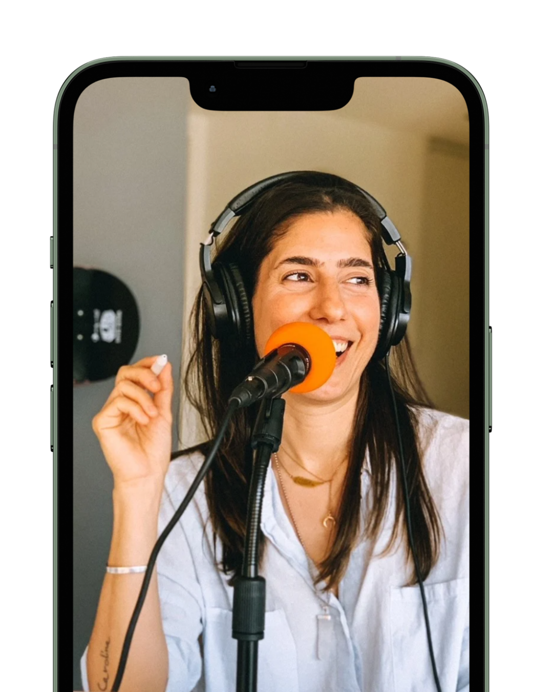 Young woman with long dark hair smiling and speaking into a microphone with an orange windscreen, wearing headphones, during a radio broadcast or podcast.