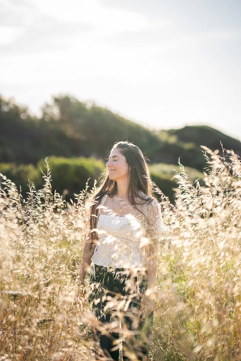 A woman walking through a field of tall, golden grass on a sunny day, with hills and trees in the background.