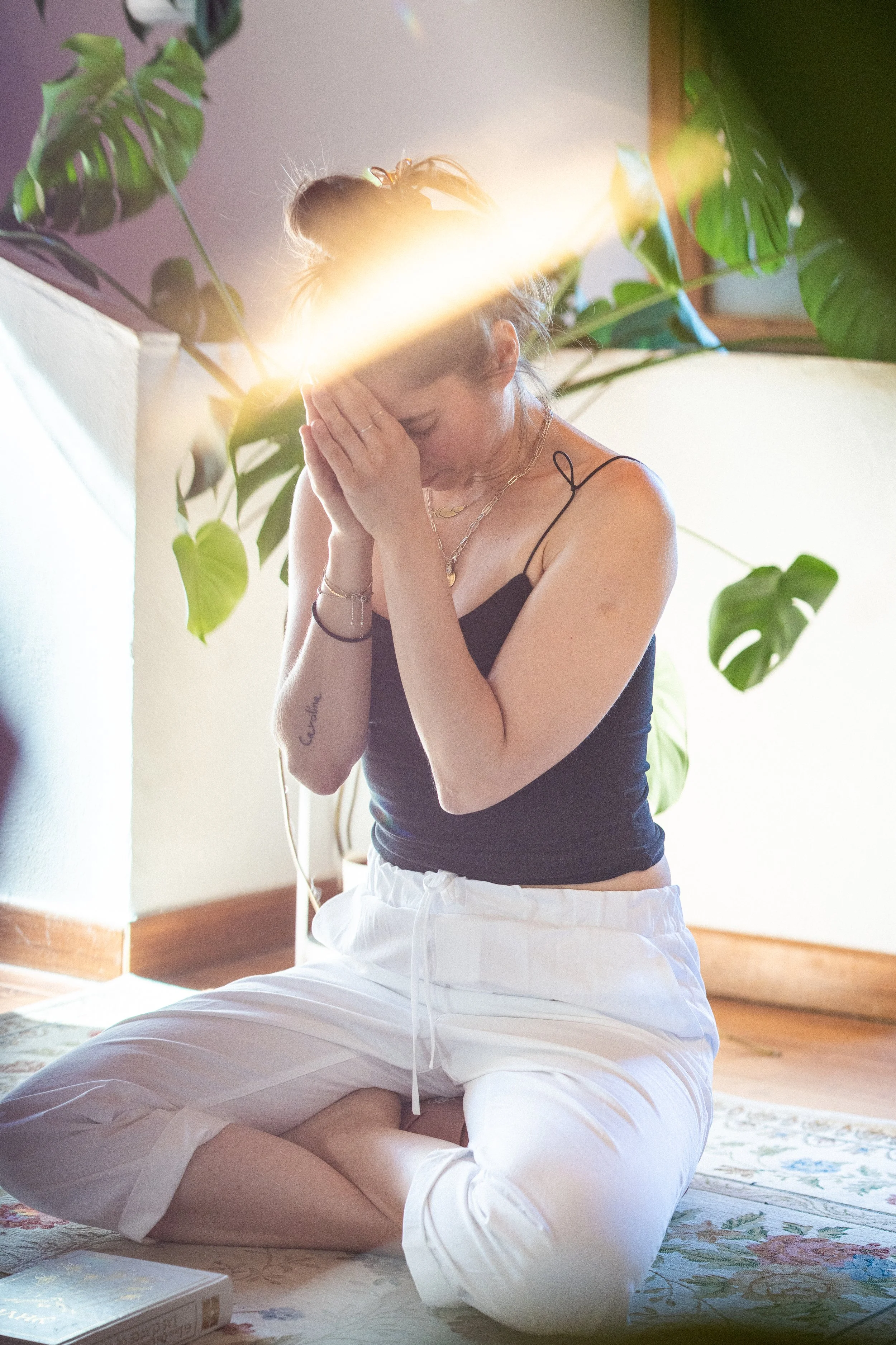 A woman sitting cross-legged on a carpet, covering her face with her hands, with sunlight coming through a window behind her, surrounded by large green plants.